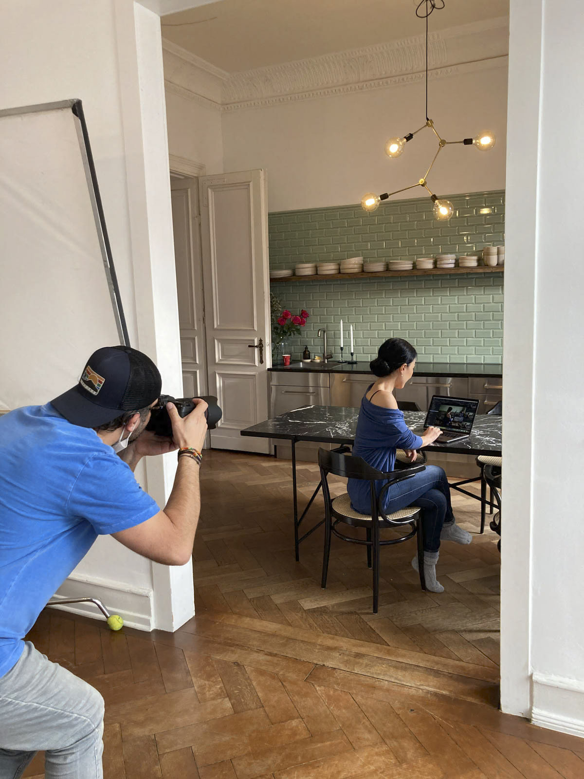 Ein Fotograf in blauem Hemd und Baseballkappe fotografiert eine Frau, die mit einem Laptop an einem Küchentisch sitzt. Die Küche ist mit grünen U-Bahn-Fliesen, einem Tisch mit Marmorplatte, Holzboden und einer modernen Leuchte ausgestattet. Die Frau schaut beim Arbeiten auf ihren Bildschirm. © Fotografie Tomas Rodriguez
