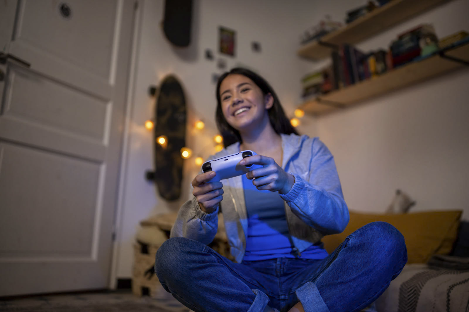 Eine Person sitzt im Schneidersitz auf einem Bett, lächelt und spielt ein Videospiel mit einem Controller. Der Raum ist gemütlich mit warmer Beleuchtung, einem Skateboard an der Wand und Regalen voller Bücher und Deko im Hintergrund. © Fotografie Tomas Rodriguez