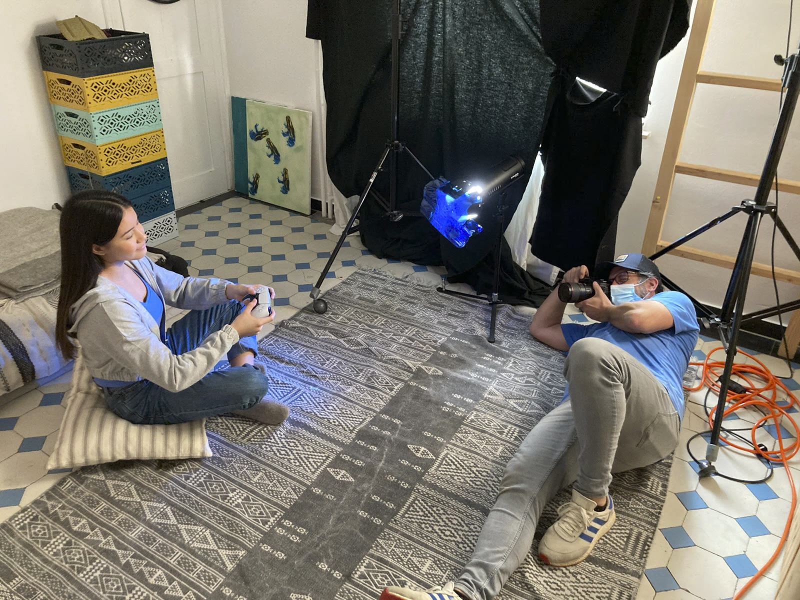 Ein Fotograf mit einer Maske liegt auf dem Boden und fotografiert eine sitzende Frau in einem Heimstudio. Die Frau hält eine weiße Tasse und sitzt auf gemusterten Kissen. Im Hintergrund sind Studioleuchten, ein Hintergrund und ein Stapel bunter Aufbewahrungskörbe zu sehen. © Fotografie Tomas Rodriguez