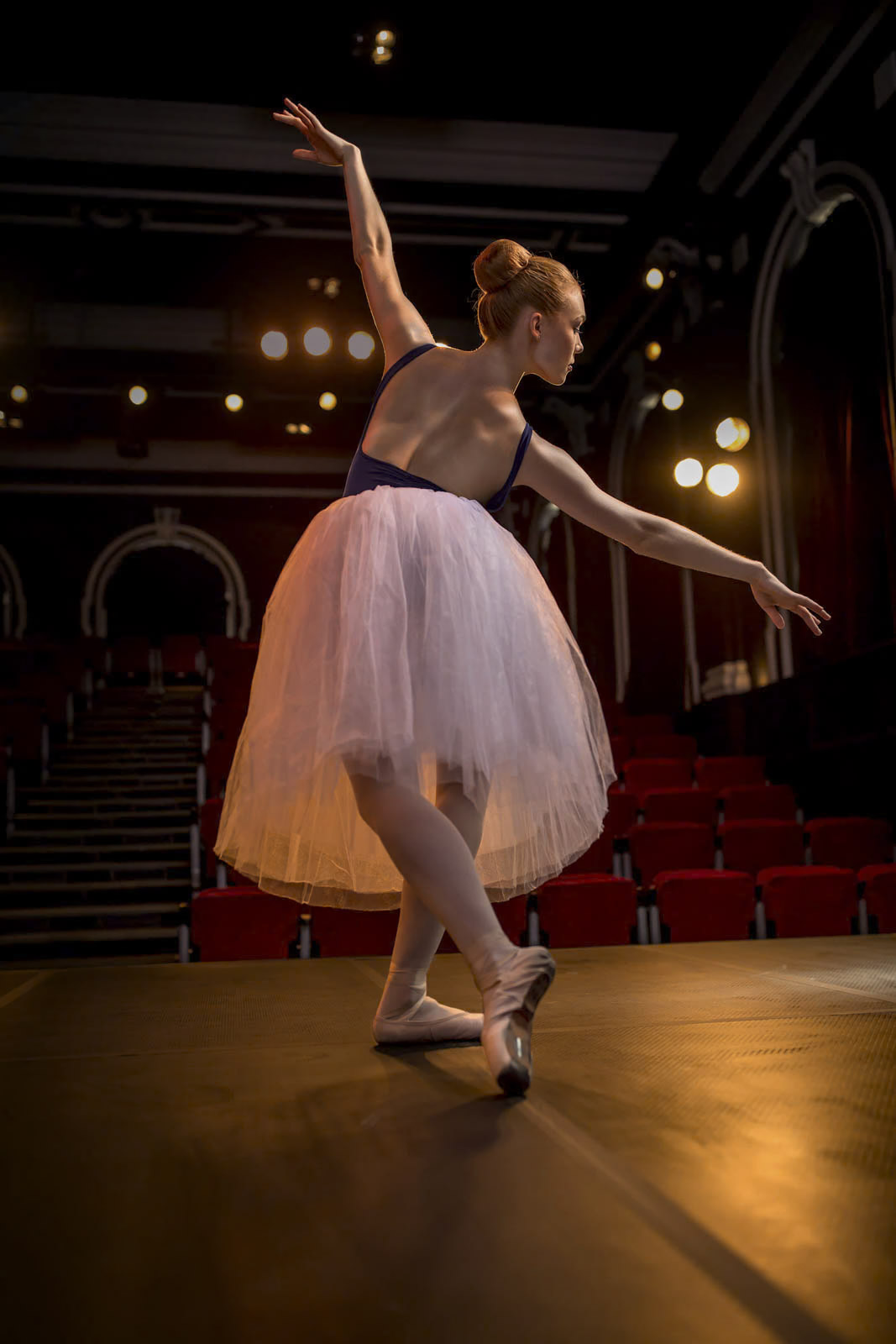 Eine Ballerina in einem weißen Tutu und einem schwarzen Trikot führt bei sanftem, warmem Licht einen anmutigen Tanz auf der Bühne auf. Das Theater ist schwach beleuchtet, im Hintergrund sind leere rote Sitze zu sehen. Die Tänzerin steht auf Spitze und streckt elegant ihre Arme aus. © Fotografie Tomas Rodriguez