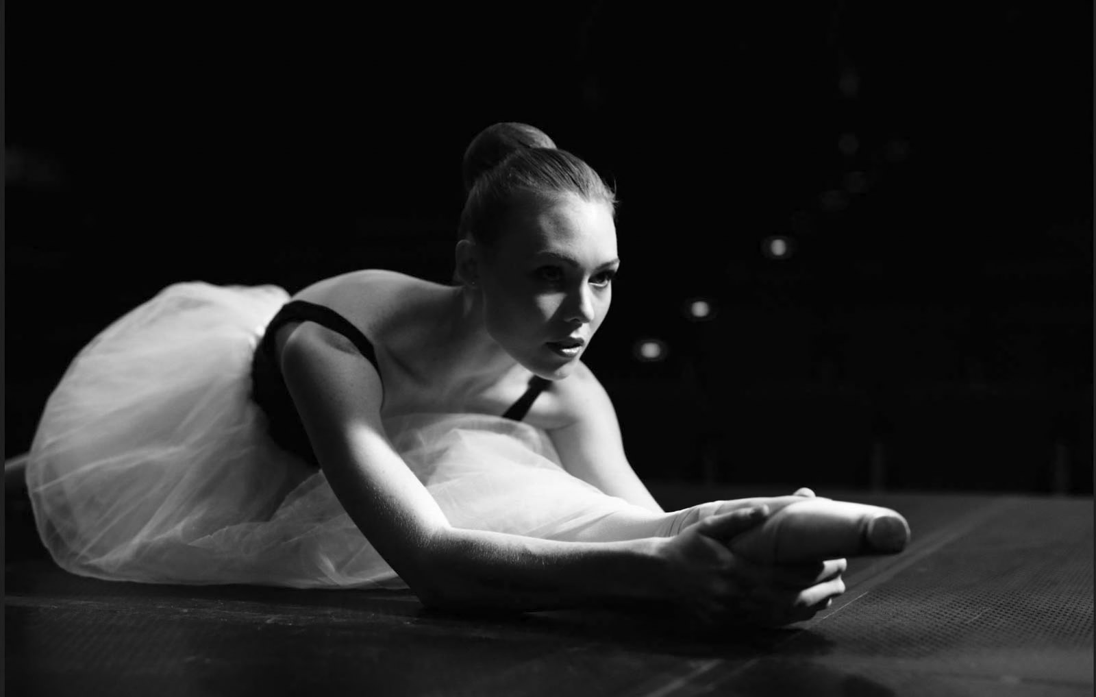 Eine Ballerina in einem Tutu streckt sich in einer schwach beleuchteten Umgebung auf dem Boden aus. Ihr Gesichtsausdruck ist konzentriert und sie streckt sich mit beiden Händen nach vorne, um ihr ausgestrecktes Bein zu halten. Das Schwarzweißbild betont die dramatische Beleuchtung und ihre anmutige Haltung. © Fotografie Tomas Rodriguez