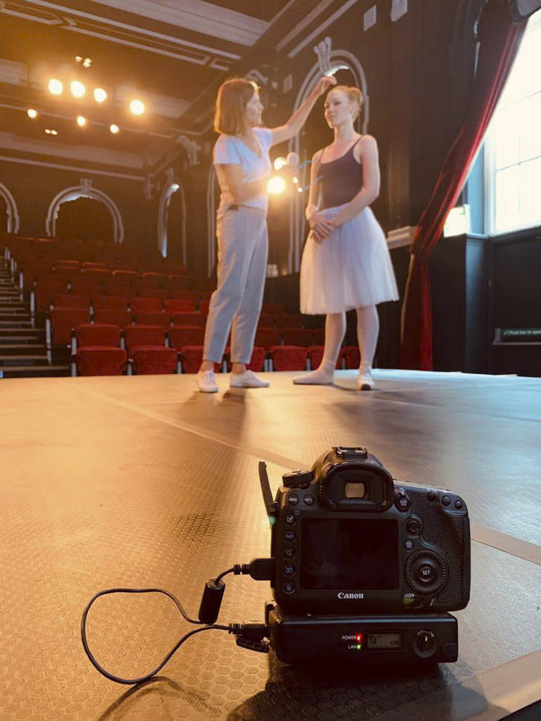 Eine Ballettlehrerin gestikuliert in Richtung einer Ballerina, die auf der Bühne eines Theaters steht. Die Ballerina trägt ein Tutu und Ballettschuhe. Im Vordergrund ist eine professionelle Canon-Kamera positioniert, die die Interaktion aufzeichnet. Im Hintergrund sind rote Sitze zu sehen. © Fotografie Tomas Rodriguez