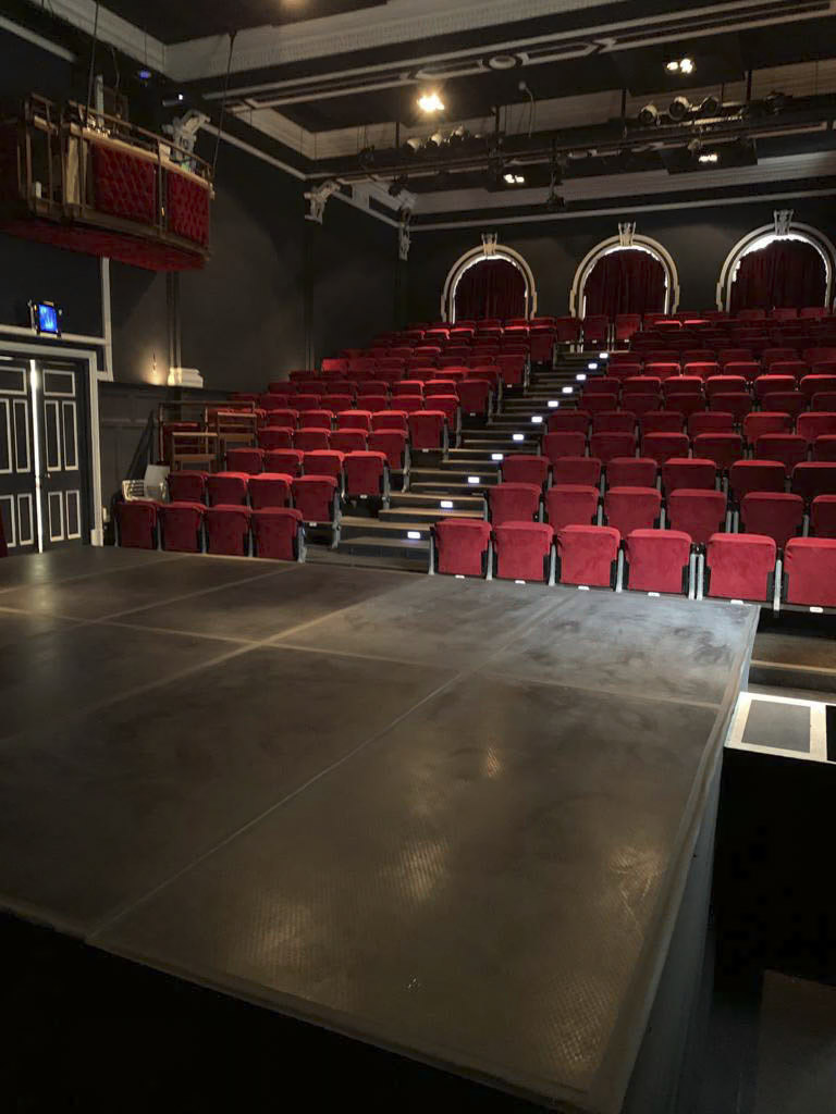 Ein schwach beleuchtetes Theater zeigt eine leere Bühne mit schwarzem Boden. Rote Samtsitze sind in Reihen mit Blick auf die Bühne angeordnet. Die hintere Reihe hat markante Bögen und kleine Lichter beleuchten die Treppen zwischen den Sitzen. Die Atmosphäre ist heiter und bereit für ein Publikum. © Fotografie Tomas Rodriguez