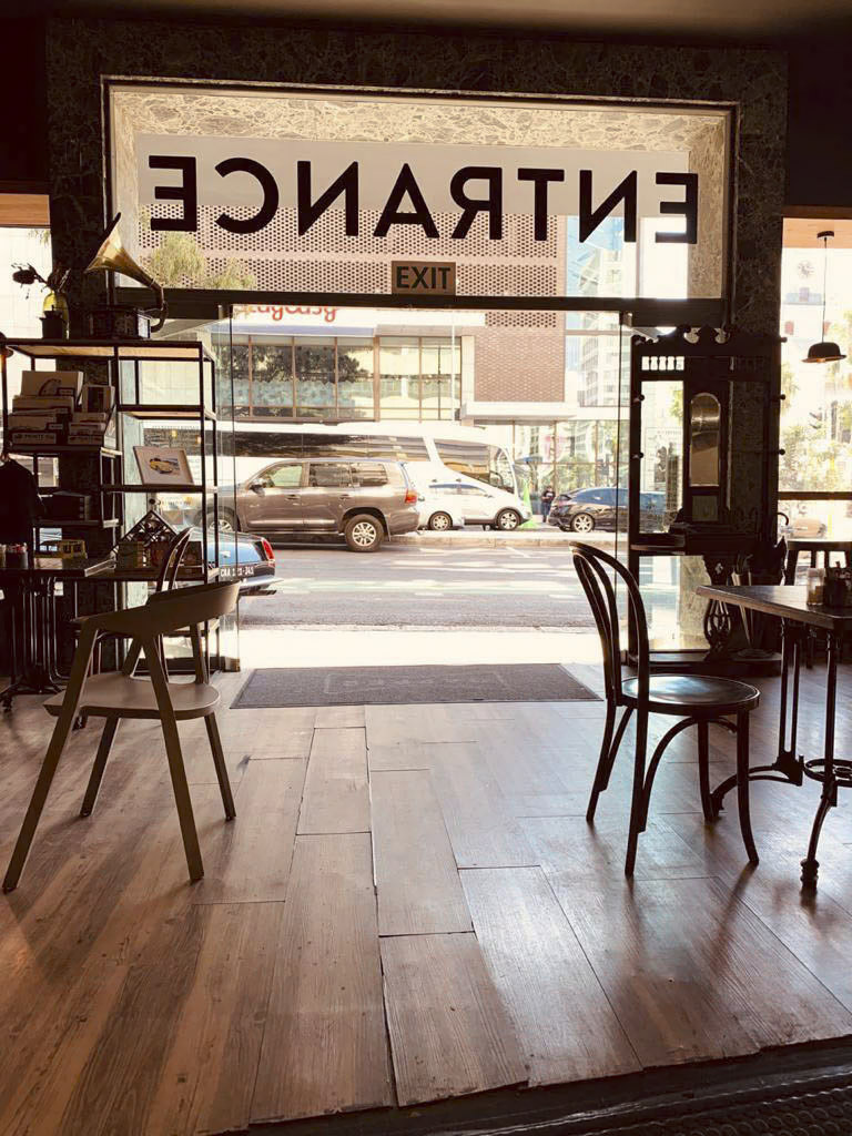 Ein gemütliches Café-Interieur mit Holzboden und ein paar Tischen und Stühlen. Die Glastür am Eingang trägt von innen gesehen ein großes, umgekehrtes „EINGANG“-Schild. Auf der Straße draußen sieht man geparkte Fahrzeuge und in Sonnenlicht getauchte Gebäude. © Fotografie Tomas Rodriguez