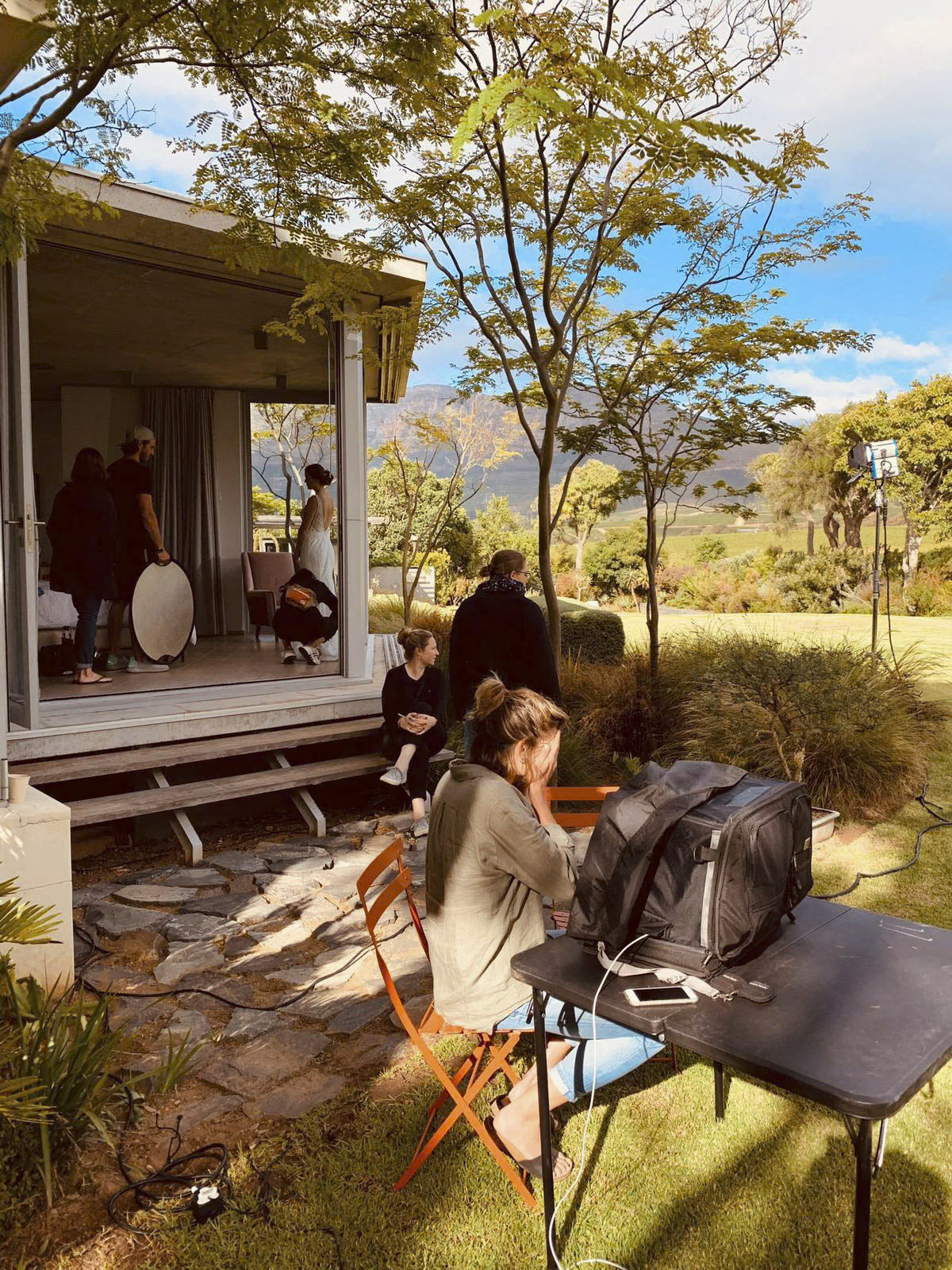 Ein Blick hinter die Kulissen eines Fotoshootings im Freien. Mehrere Personen haben sich um eine Veranda versammelt, einige bereiten die Ausrüstung vor, andere richten einen Hintergrund aus. Eine Person sitzt mit einer Kameratasche an einem Tisch in der Nähe, umgeben von üppigem Grün und einer Berglandschaft. © Fotografie Tomas Rodriguez