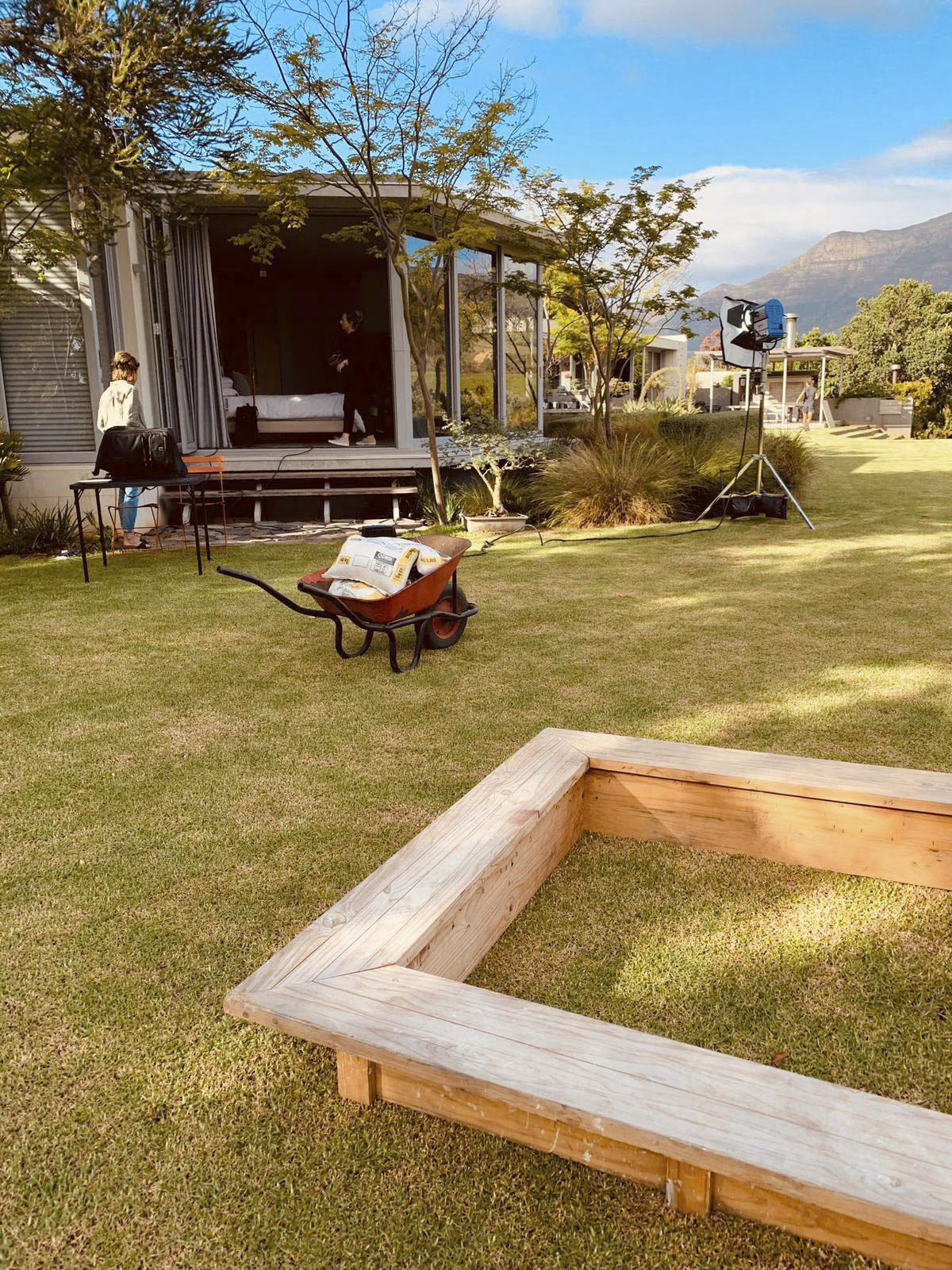 Eine malerische Außenkulisse mit einem modernen Haus im Hintergrund und Bergen in der Ferne. Eine Schubkarre voller Säcke steht auf dem Gras neben einem Holzrahmen. Zwei Personen stehen auf der Terrasse des Hauses im Schatten der Bäume in der Nähe. © Fotografie Tomas Rodriguez