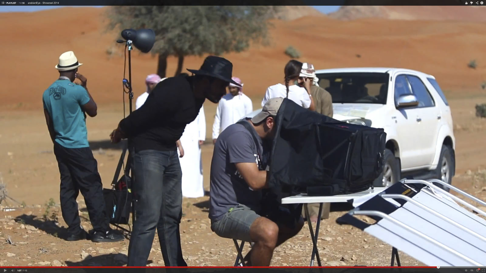 Filmteam bei der Arbeit in einer Wüstenumgebung. Eine Person in grauem Hemd und Mütze bedient eine Kamera unter einer schwarzen Abdeckung, während eine andere Person neben ihr ein schwarzes Hemd und eine schwarze Mütze trägt. Mehrere Personen mit Hüten und traditioneller Kleidung stehen im Hintergrund neben einem weißen SUV. © Fotografie Tomas Rodriguez