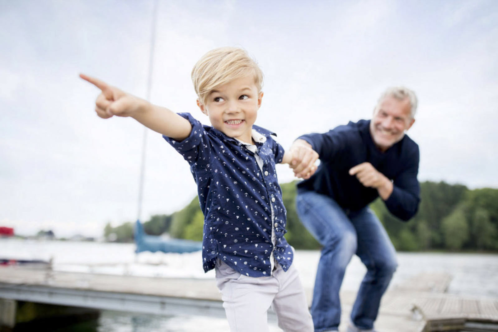 Ein kleiner Junge mit blondem Haar und einem blauen gepunkteten Hemd zeigt aufgeregt zur Seite, während er auf einem Dock steht. Er hält die Hand eines älteren Mannes in einem marineblauen Pullover, der lächelt und in die gleiche Richtung gestikuliert. Im Hintergrund sind ein Segelboot und Bäume zu sehen. © Fotografie Tomas Rodriguez