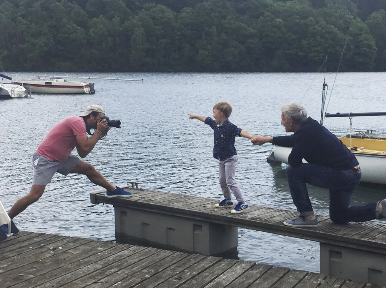 Ein Mann in rosa Hemd und Shorts macht auf einem Holzsteg neben einem See ein Foto mit einer Kamera. Ein kleines Kind steht auf dem Steg und zeigt in die Ferne, während ein anderer Mann niederkniet und die Hand des Kindes hält. Im Hintergrund sind Bäume und Boote zu sehen. © Fotografie Tomas Rodriguez