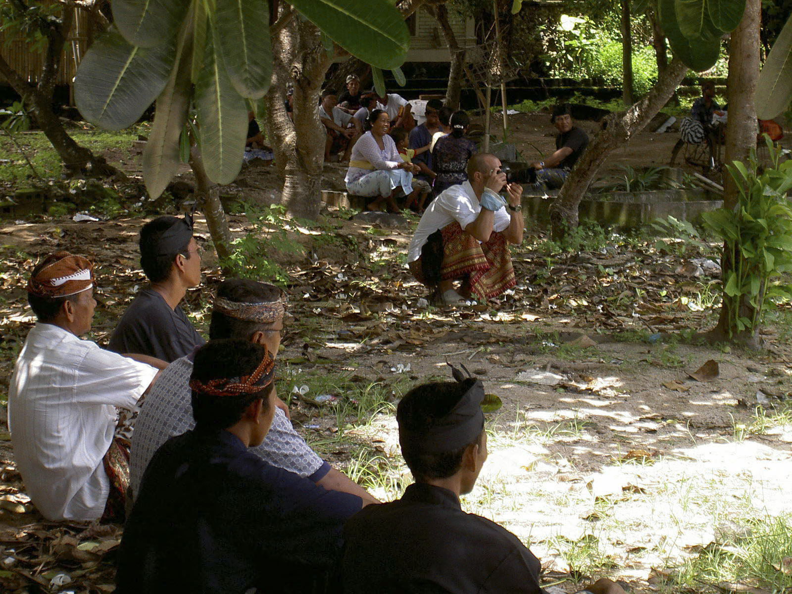 Eine Gruppe von Menschen sitzt in traditioneller Kleidung und Kopfbedeckungen im Schatten von Bäumen in einem Waldgebiet auf dem Boden. Einige sitzen im Gras, andere auf Felsen, und eine Person hockt mit einer Kamera in der Nähe. Verschiedene Gegenstände liegen verstreut herum. © Fotografie Tomas Rodriguez