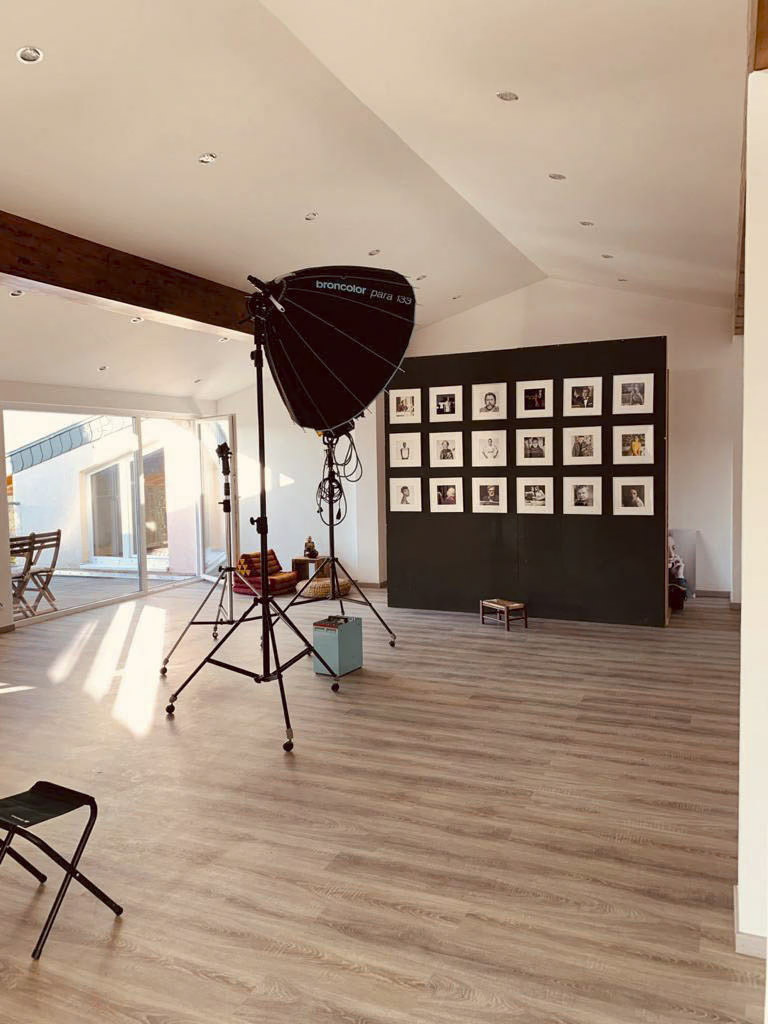 Ein geräumiges Fotostudio mit Holzboden verfügt über eine große Softbox-Leuchte auf einem Ständer, einen Klappstuhl und eine Wand mit gerahmten Schwarzweißfotos. Sonnenlicht strömt durch große Fenster herein und erhellt den Raum teilweise. © Fotografie Tomas Rodriguez