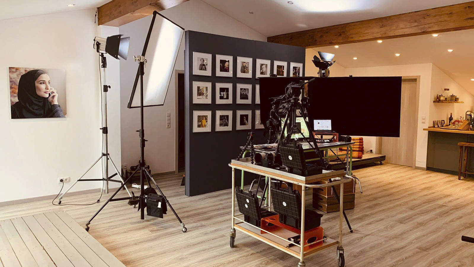 Ein Fotostudio mit Beleuchtungsausrüstung, Kameras auf Stativen und einem Monitor auf einem Rollwagen. Der Raum hat einen Holzboden, eine Balkendecke und eine mit gerahmten Fotos geschmückte Wand. An einer separaten Wand hängt das Porträt einer Frau. © Fotografie Tomas Rodriguez