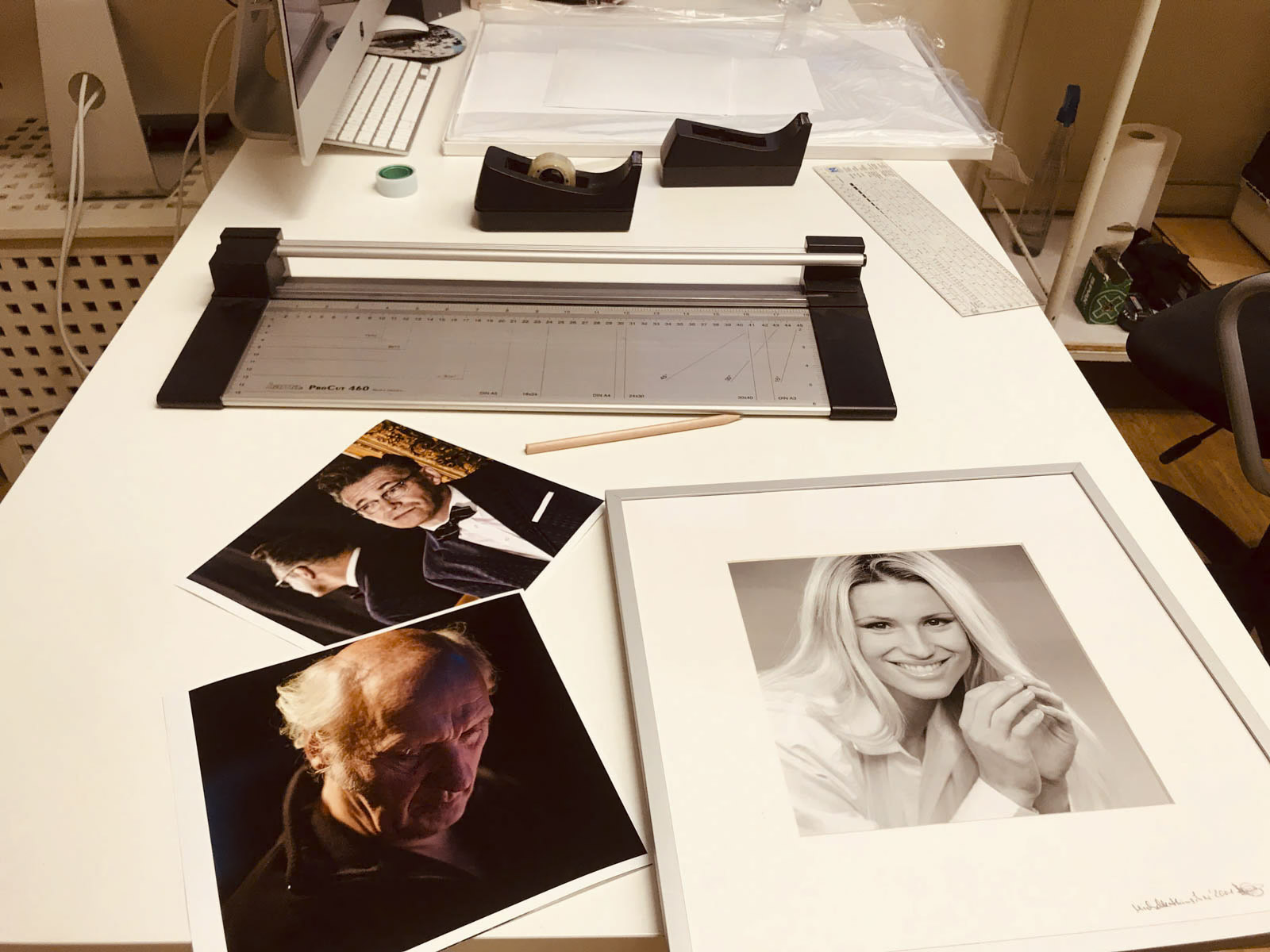 Ein weißer Schreibtisch mit einem großen Papierschneider, zwei losen Fotos von Menschen, einem gerahmten Schwarzweißporträt einer Frau, Papierkram, einem Bleistift und anderen Büromaterialien. Mehrere Gegenstände liegen verstreut herum, was darauf hindeutet, dass an dem Schreibtisch gearbeitet wird. © Fotografie Tomas Rodriguez
