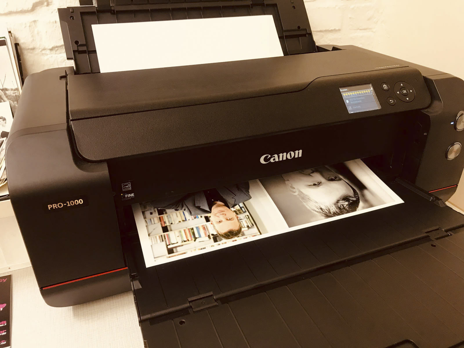 Ein Canon PRO-1000-Drucker druckt gerade ein Blatt mit zwei Schwarzweiß-Porträtfotos. Der Drucker hat ein elegantes schwarzes Design mit einem kleinen Bildschirm und Tasten auf der rechten Seite. Der Hintergrund besteht aus einer Ziegelsteinmauer und einer weißen Oberfläche. © Fotografie Tomas Rodriguez