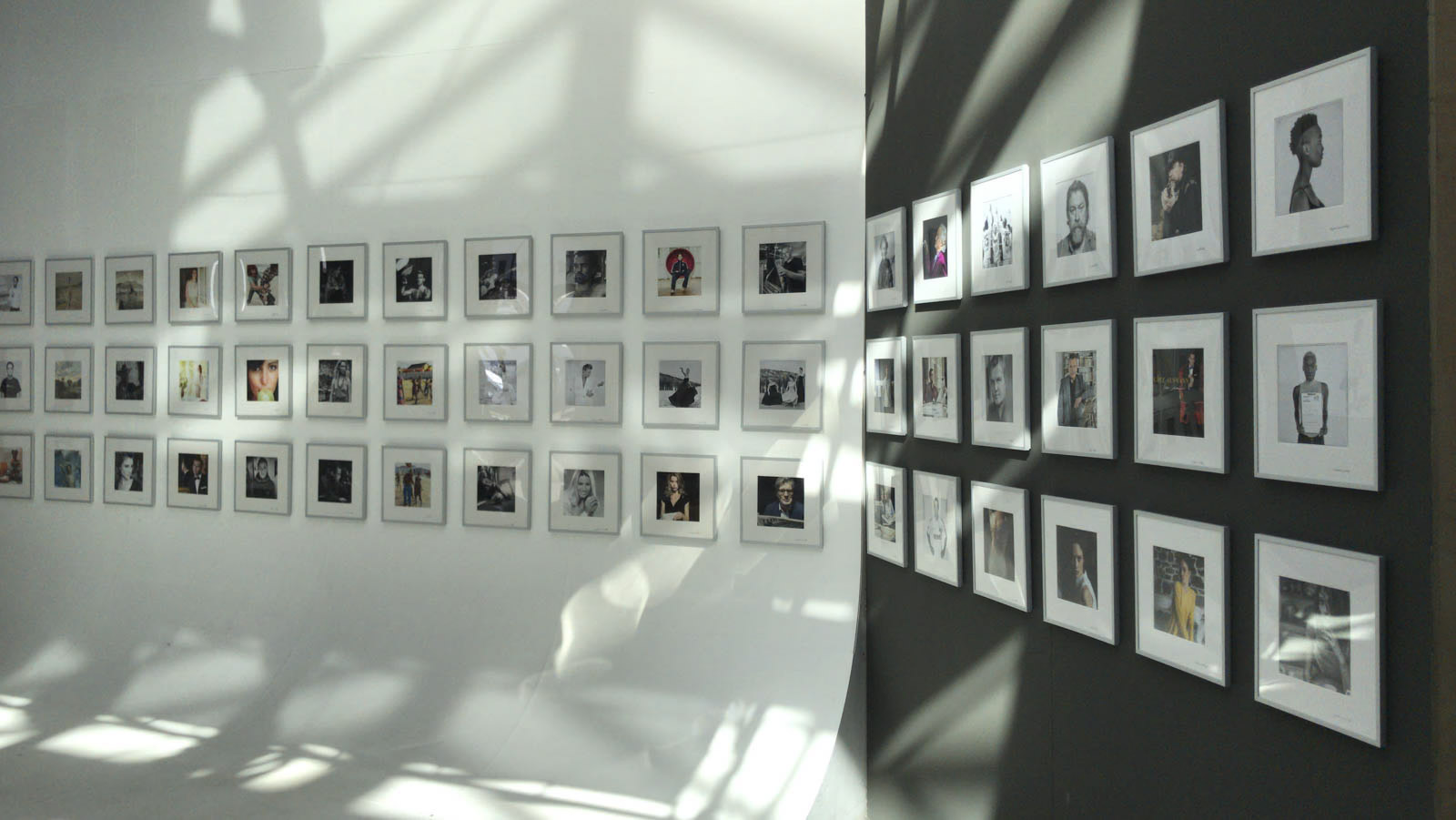 Eine Fotogalerie mit mehreren gerahmten Bildern, die in Reihen an weißen und dunklen Wänden ausgestellt sind. Schatten der Deckenbeleuchtung erzeugen Muster an den Wänden und auf dem Boden und verleihen dem Raum Tiefe und Struktur. Die Bilder haben unterschiedliche Inhalte und sorgen so für visuelles Interesse. © Fotografie Tomas Rodriguez
