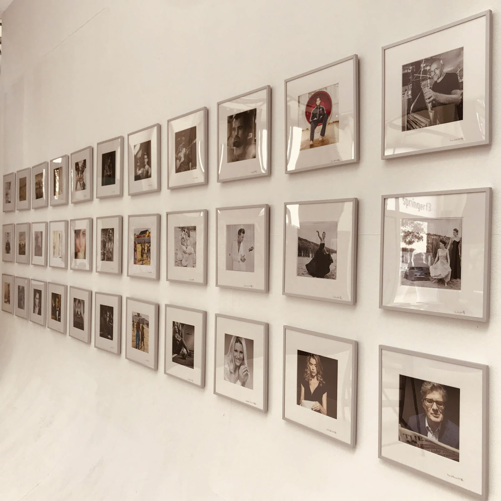 Eine Wand, die mit einem ordentlich angeordneten Raster aus gerahmten Schwarzweißfotos geschmückt ist. Jeder Rahmen enthält ein anderes Porträt, wobei in drei Reihen insgesamt vierundzwanzig Bilder angezeigt werden. Die Rahmen werfen subtile Schatten und verleihen der Ausstellung Dimension. © Fotografie Tomas Rodriguez