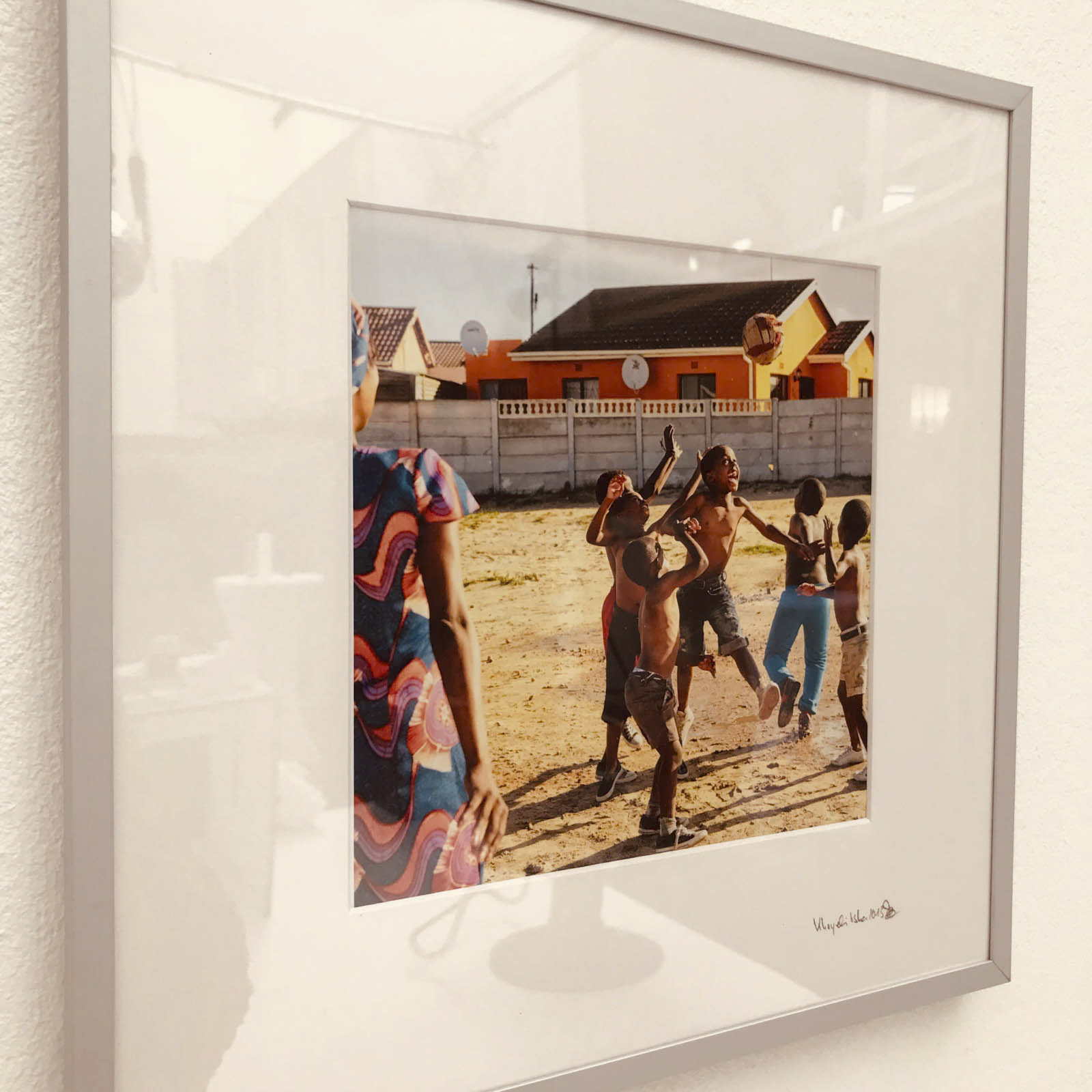 Ein gerahmtes Foto, das an einer weißen Wand hängt. Das Bild zeigt mehrere Kinder, die draußen spielen, wobei einige Kinder mitten in der Bewegung sind, möglicherweise springen oder nach etwas in der Luft greifen. Im Hintergrund sind bunte Gebäude und ein Zaun zu sehen. © Fotografie Tomas Rodriguez