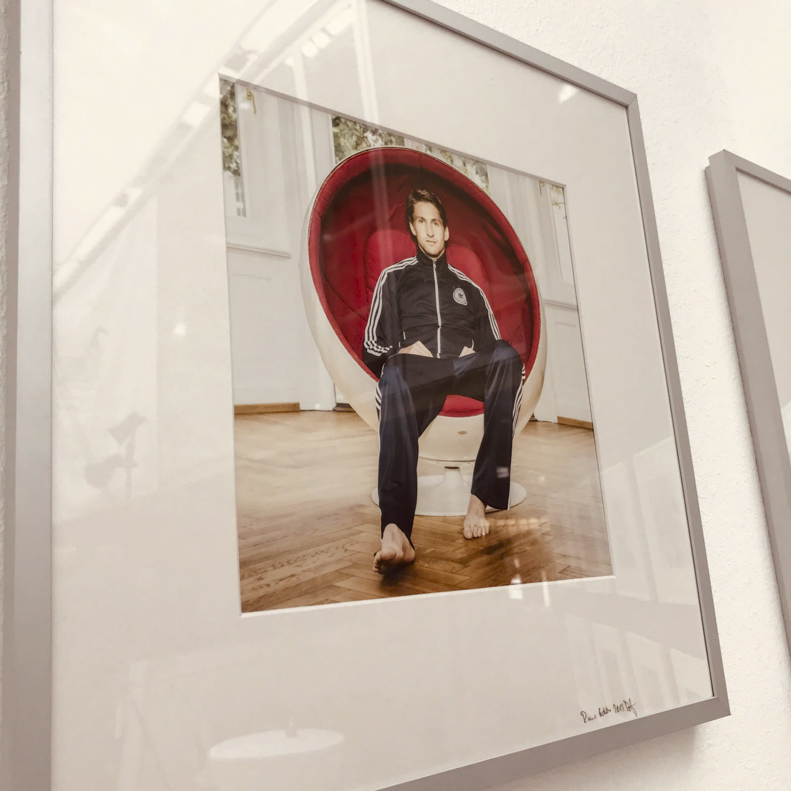 Ein gerahmtes Foto an einer weißen Wand zeigt eine Person, die barfuß in einem roten, runden Stuhl sitzt. Die Person trägt eine dunkle Trainingsjacke und eine Hose mit weißen Streifen. Der Fußboden ist aus Holz, im Hintergrund sind Fenster mit weißen Vorhängen zu sehen. © Fotografie Tomas Rodriguez