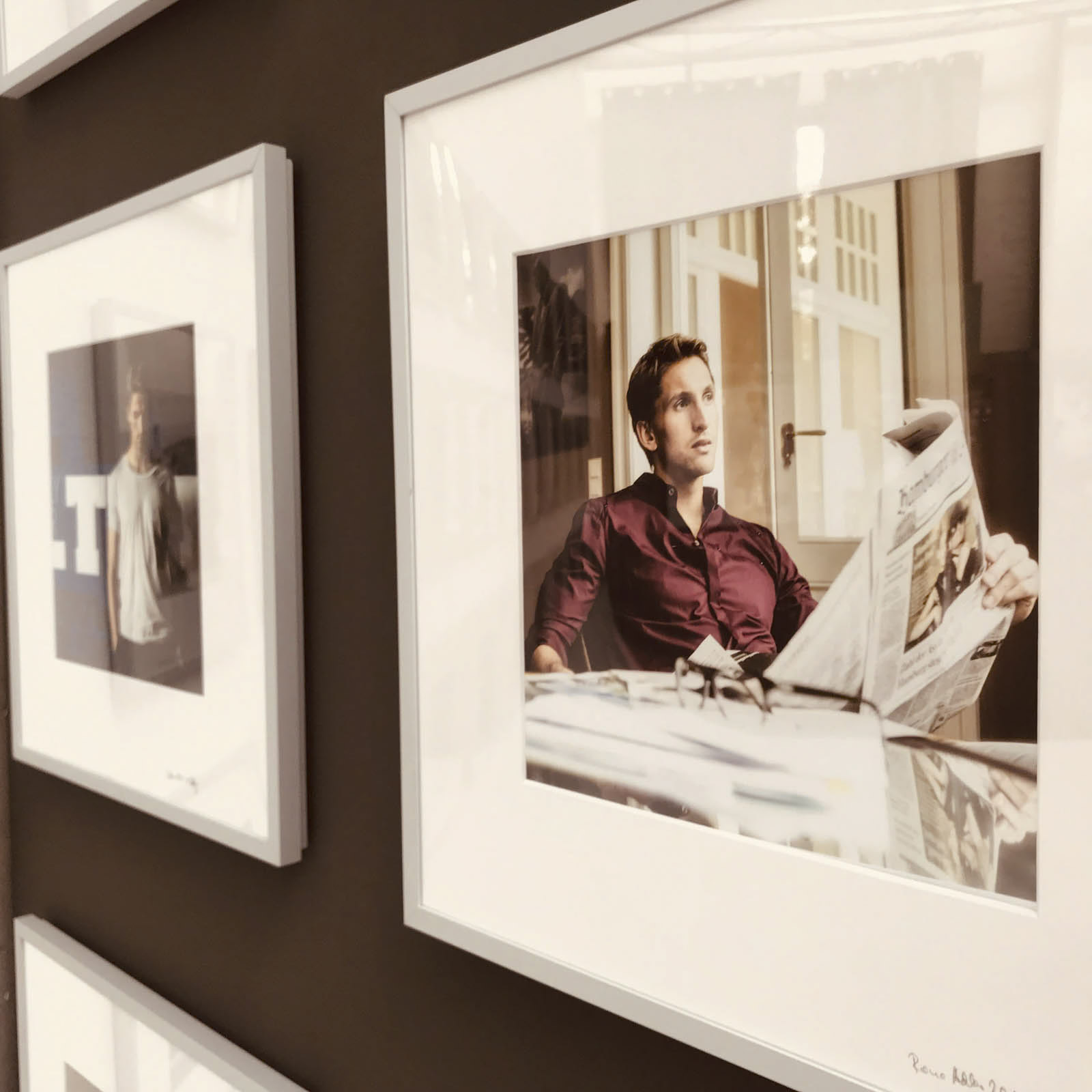 Gerahmte Fotos an einer Wand. Das prominente Foto zeigt eine Person in einem kastanienbraunen Hemd, die an einem Schreibtisch eine Zeitung liest. Daneben hängt ein weiteres Foto, das eine Person in Freizeitkleidung zeigt, die vor einem Hintergrund mit dem Buchstaben „T“ steht. © Fotografie Tomas Rodriguez