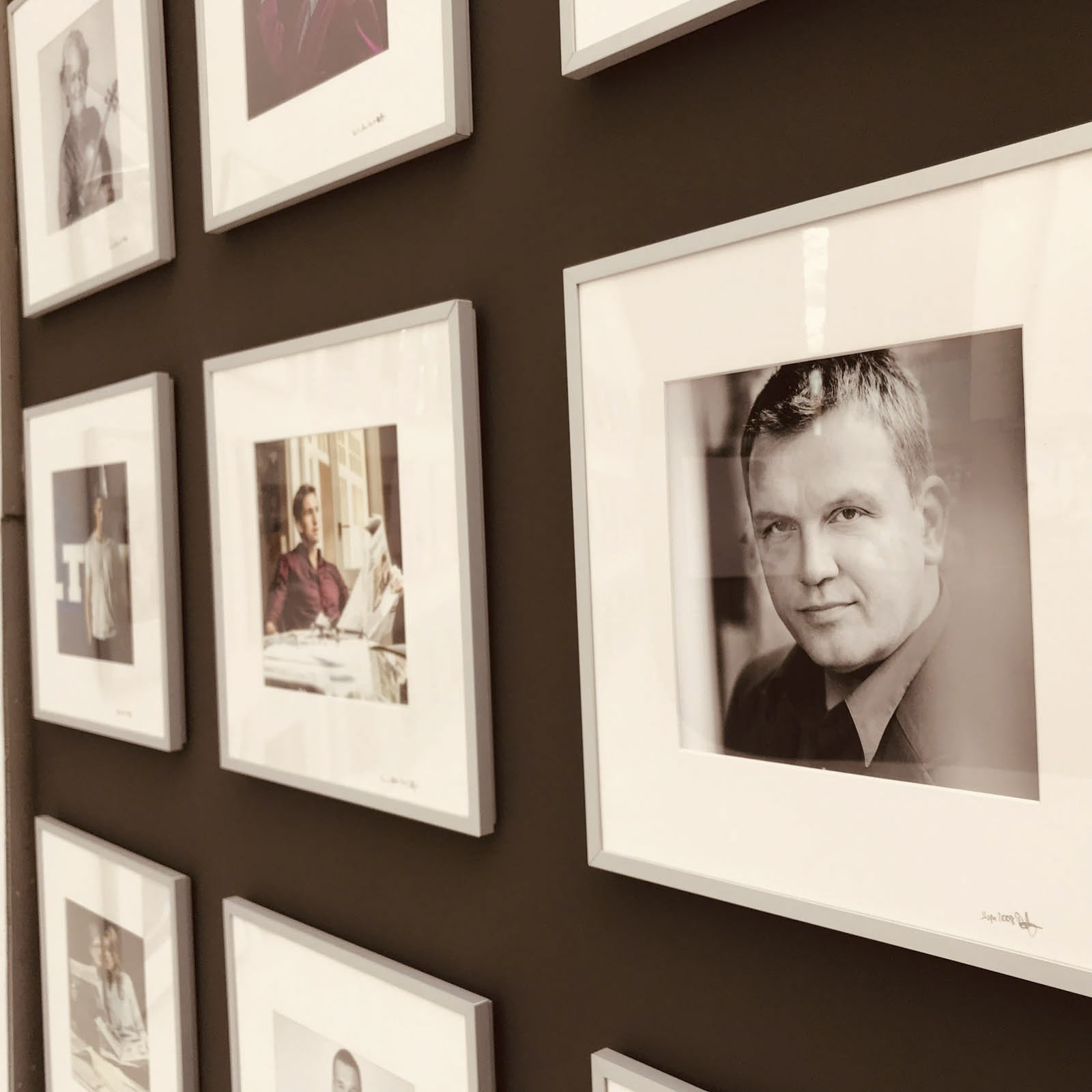 Eine Wand, die mit einem Raster aus gerahmten Schwarzweißfotos dekoriert ist. Jeder Rahmen enthält ein Porträt einer Person. Das Bild konzentriert sich auf ein bestimmtes Porträt eines Mannes im Vordergrund, während die anderen Porträts im Hintergrund leicht unscharf sind. © Fotografie Tomas Rodriguez