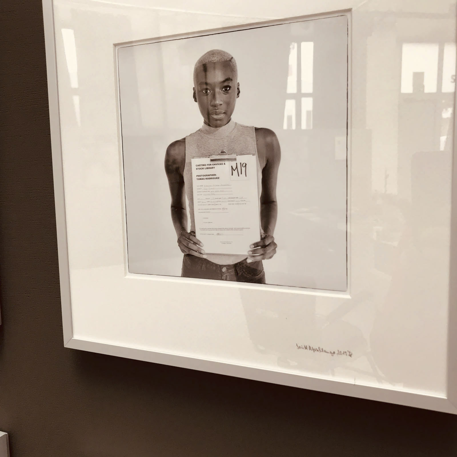 Ein gerahmtes Foto einer Person mit kurzen Haaren, die ein Blatt Papier hält, auf dem „M19“ steht. Die Person trägt ein ärmelloses Oberteil und steht vor einem hellen Hintergrund. Der Rahmen hängt an einer Wand. © Fotografie Tomas Rodriguez