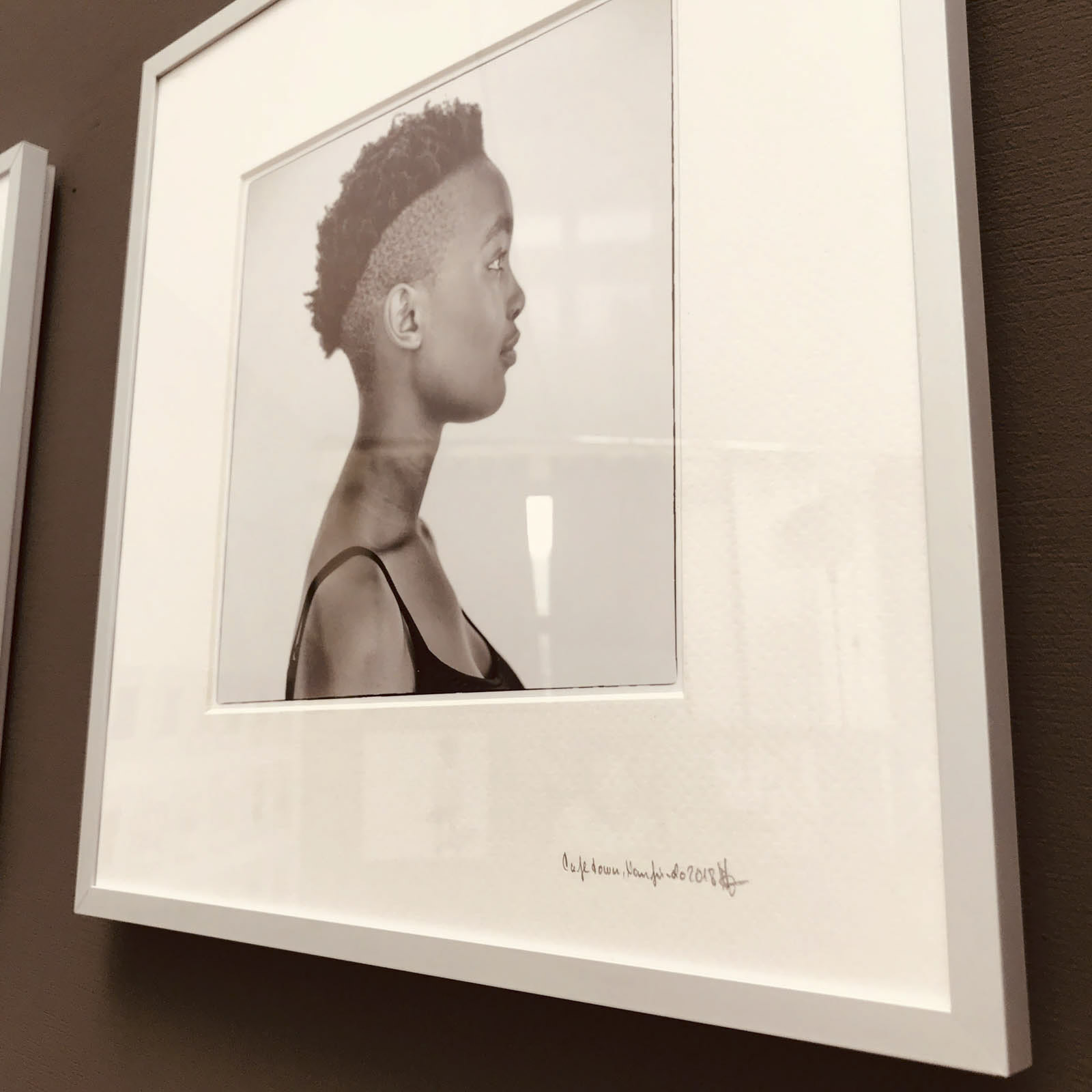 An einer Wand hängt ein gerahmtes Schwarzweiß-Porträtfoto einer Person im Profil. Die Person hat eine kurze, strukturierte Frisur und trägt ein ärmelloses Oberteil. Unter dem Foto befindet sich eine Signatur. © Fotografie Tomas Rodriguez