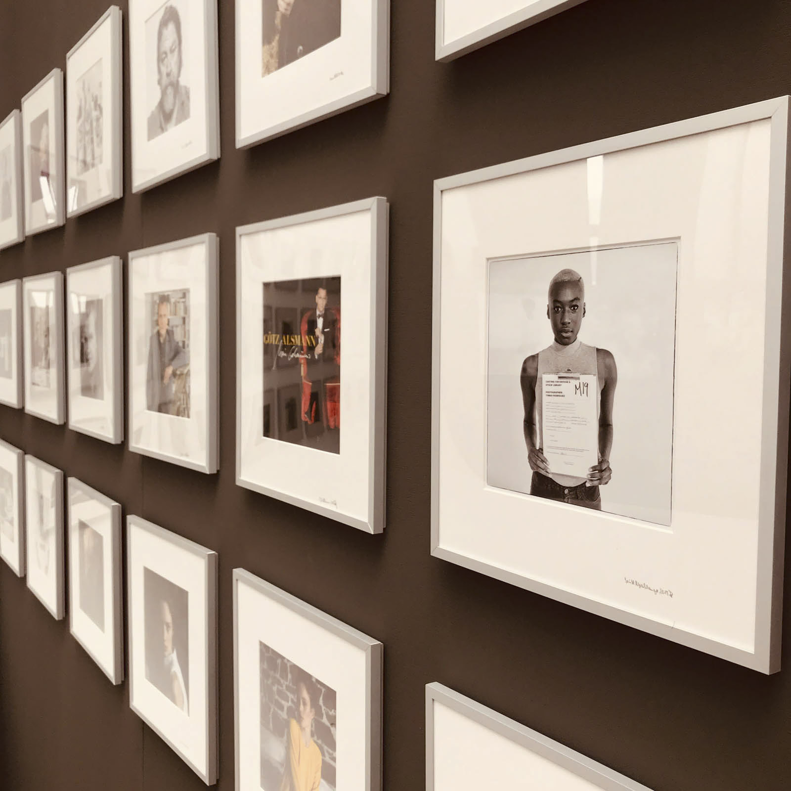 Eine braune Wand mit einer Reihe gerahmter Fotos. Die Bilder zeigen verschiedene Personen, wobei ein prominentes Bild eine Person zeigt, die ein Dokument hält. Die Sammlung scheint eine kuratierte Fotogalerie oder Ausstellung zu sein. © Fotografie Tomas Rodriguez