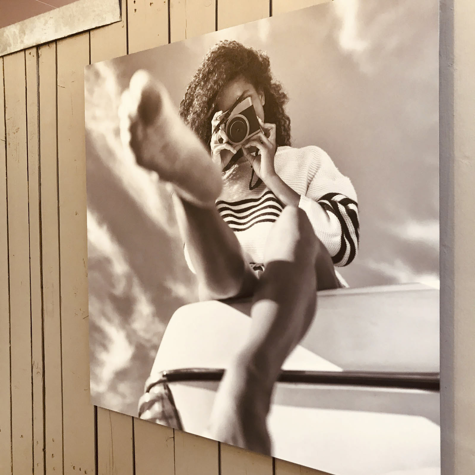 An einer Wand hängt ein Schwarzweißfoto, das eine Person in einem gestreiften Pullover zeigt, die mit einer Vintage-Kamera ein Foto macht. Das Foto wurde aus einem niedrigen Winkel aufgenommen, wobei der Fokus auf den Füßen der Person im Vordergrund liegt und darüber teilweise der Himmel und die Wolken zu sehen sind. © Fotografie Tomas Rodriguez