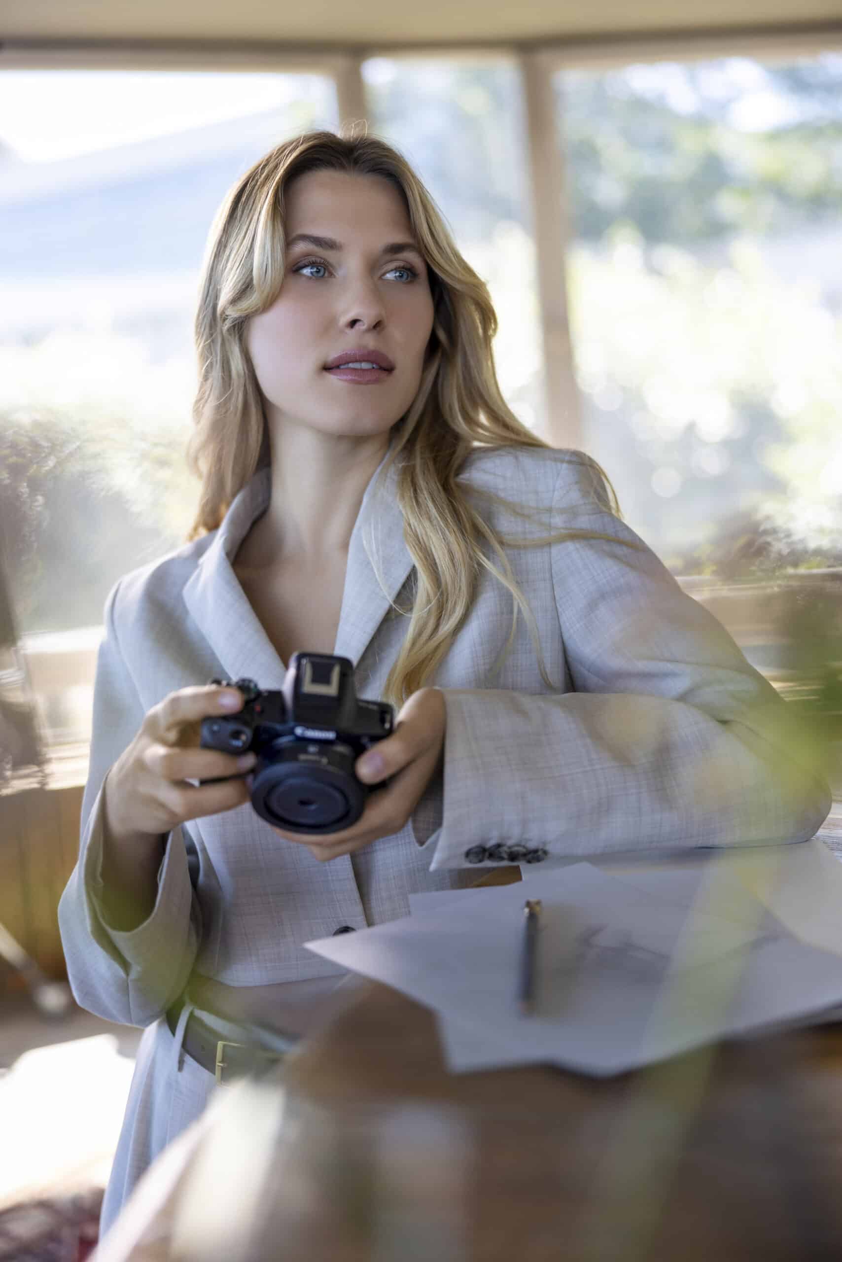 Eine Frau mit langen blonden Haaren und einem hellgrauen Blazer hält eine Kamera und blickt zur Seite. Sie sitzt an einem Tisch und hat ein paar Papiere und einen Stift vor sich. Im Hintergrund sind große Fenster zu sehen, durch die das Sonnenlicht fällt. © Fotografie Tomas Rodriguez