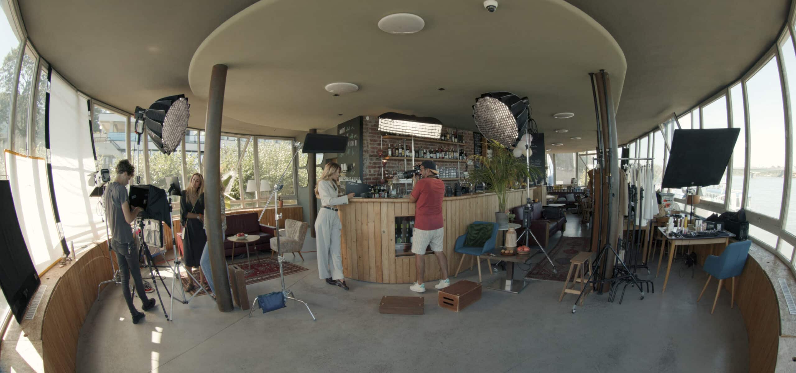 Ein modernes Indoor-Filmset mit großen Fenstern rundherum. Mehrere Personen sind damit beschäftigt, Kameras, Beleuchtung und andere Ausrüstung einzurichten. In der Mitte befindet sich ein Barbereich und im ganzen Raum sind Möbel verteilt. © Fotografie Tomas Rodriguez
