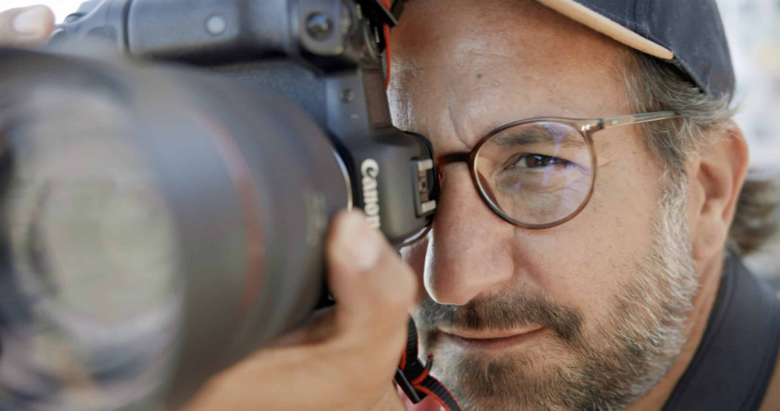 Nahaufnahme eines Mannes mit Brille und Hut, der durch den Sucher einer Canon-Kamera genau fokussiert. Sein Gesichtsausdruck ist eindringlich und konzentriert, als er eine Aufnahme macht. Der Hintergrund ist verschwommen, was die Aufmerksamkeit auf seine detaillierten Gesichtszüge und die Kamera lenkt. © Fotografie Tomas Rodriguez