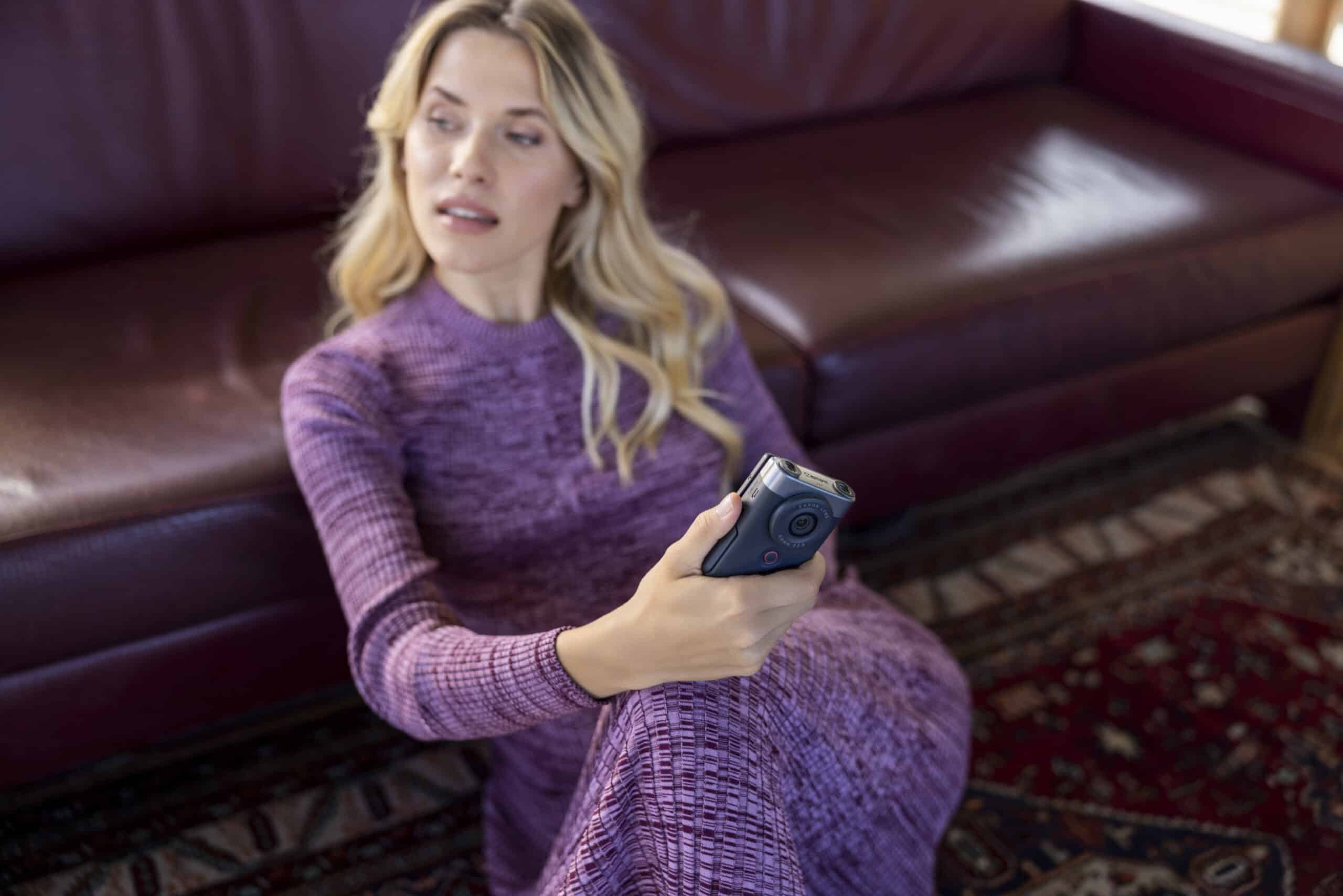 Eine Frau mit langen blonden Haaren und einem lilafarbenen Kleid sitzt auf dem Boden vor einem kastanienbraunen Ledersofa, hält eine schwarze Kamera in der Hand und blickt in sie hinein. Der Boden ist mit einem kunstvoll gemusterten Teppich bedeckt. © Fotografie Tomas Rodriguez