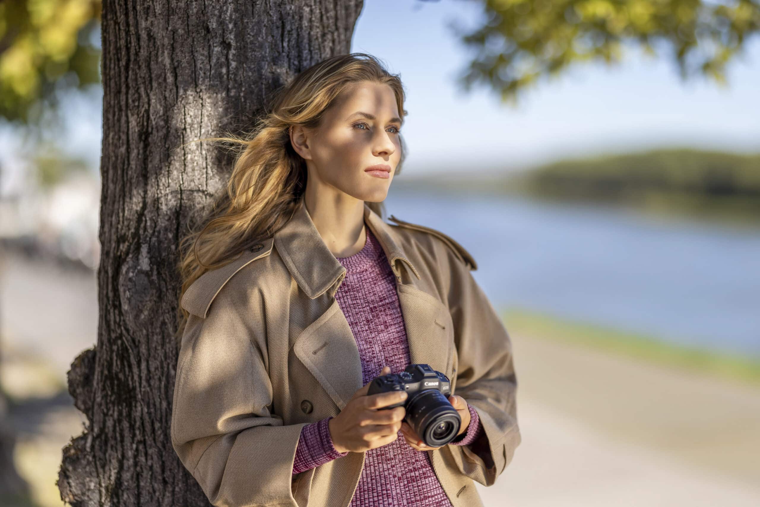Eine Frau mit langen blonden Haaren steht im Freien und lehnt an einem Baum. Sie hält eine Kamera in der Hand und trägt einen beigen Trenchcoat über einem rosa Pullover. Der Hintergrund zeigt eine ruhige Flusslandschaft mit grünem Laub und einem klaren, blauen Himmel. © Fotografie Tomas Rodriguez