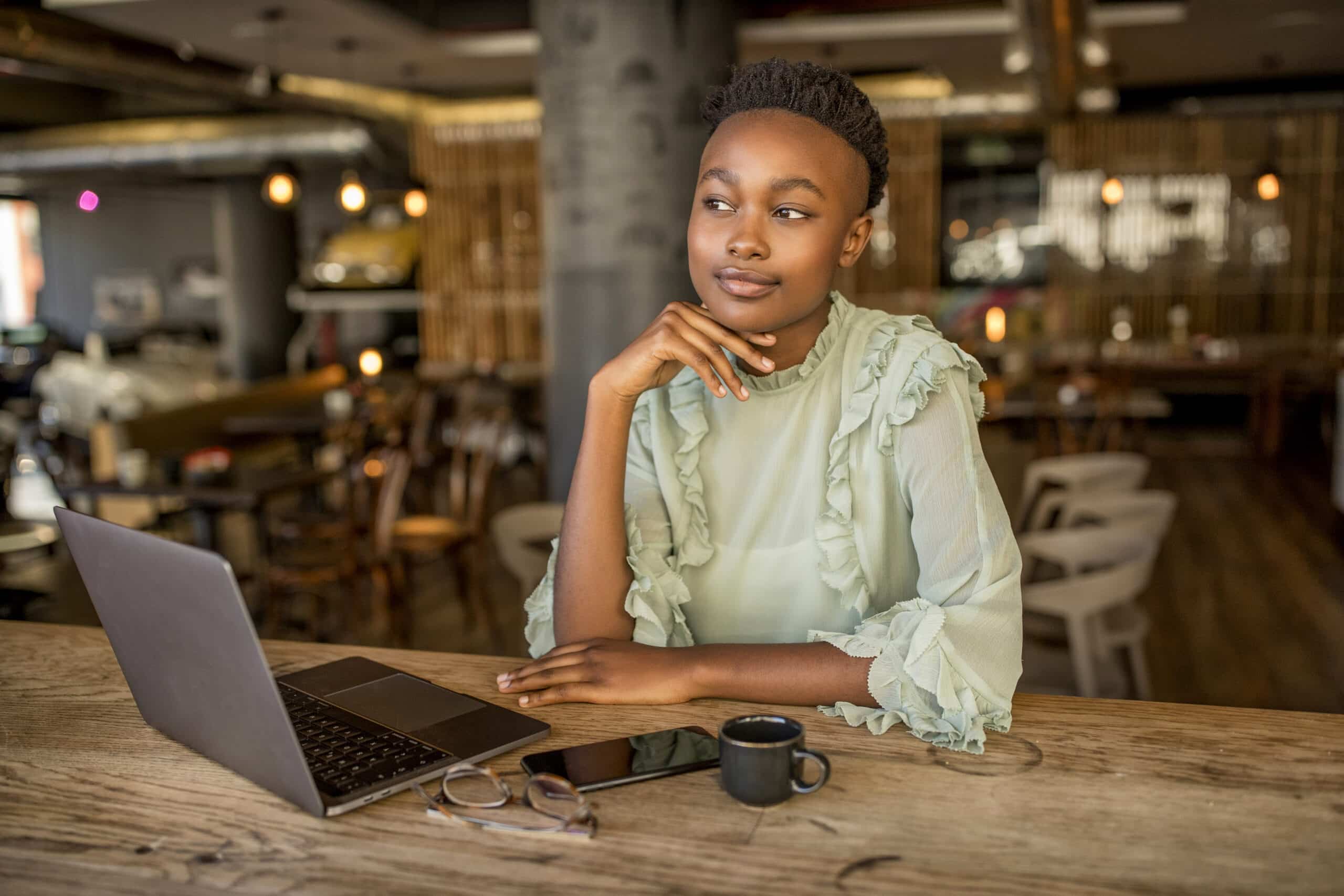 Eine junge Frau sitzt an einem Holztisch in einem modernen Café, die Hand am Kinn, und blickt nachdenklich von ihrem Laptop weg, neben dem eine kleine Tasse Kaffee und Gläser stehen. © Fotografie Tomas Rodriguez