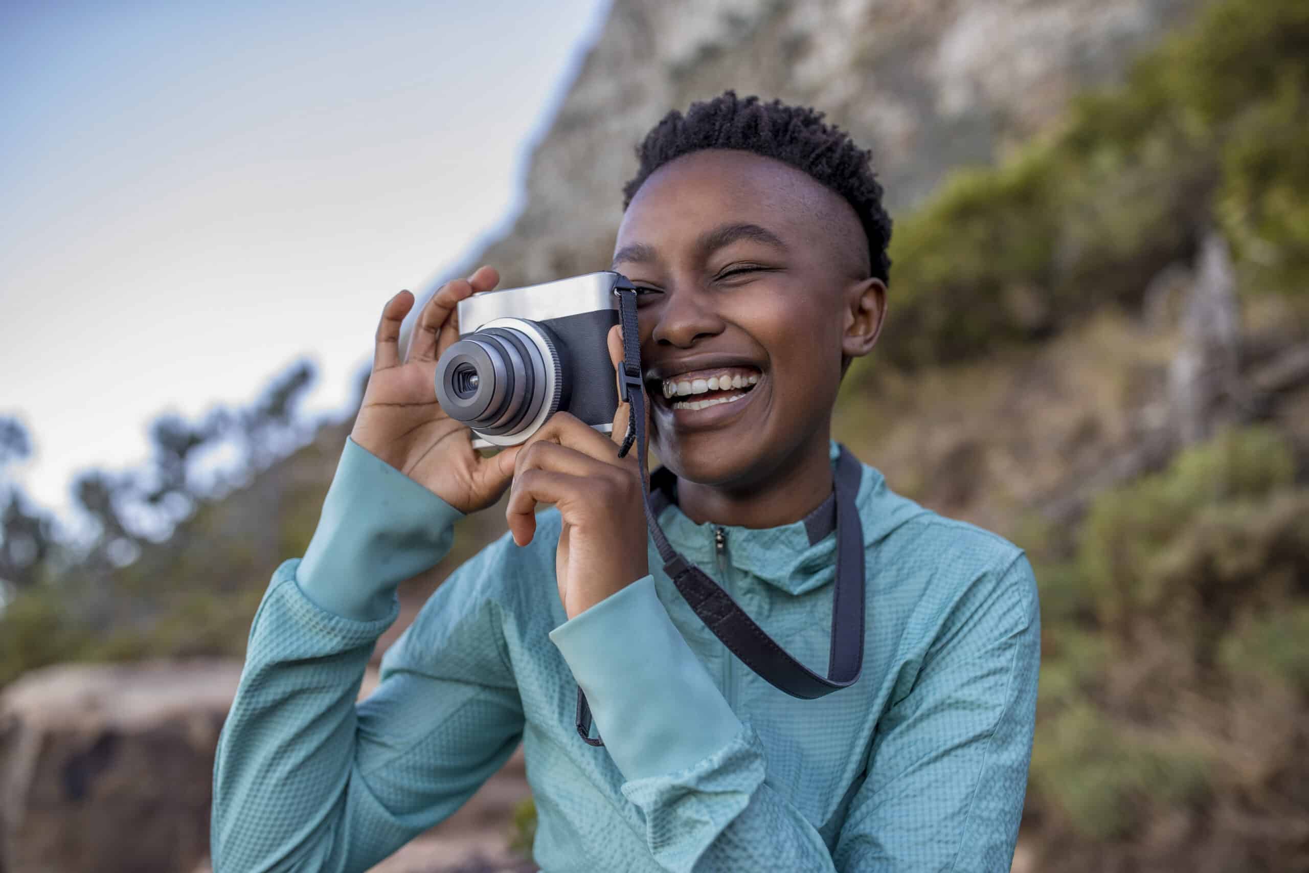 Eine fröhliche junge Frau macht mit einer Digitalkamera im Freien Fotos und lacht, während sie von der Naturlandschaft umgeben ist. © Fotografie Tomas Rodriguez