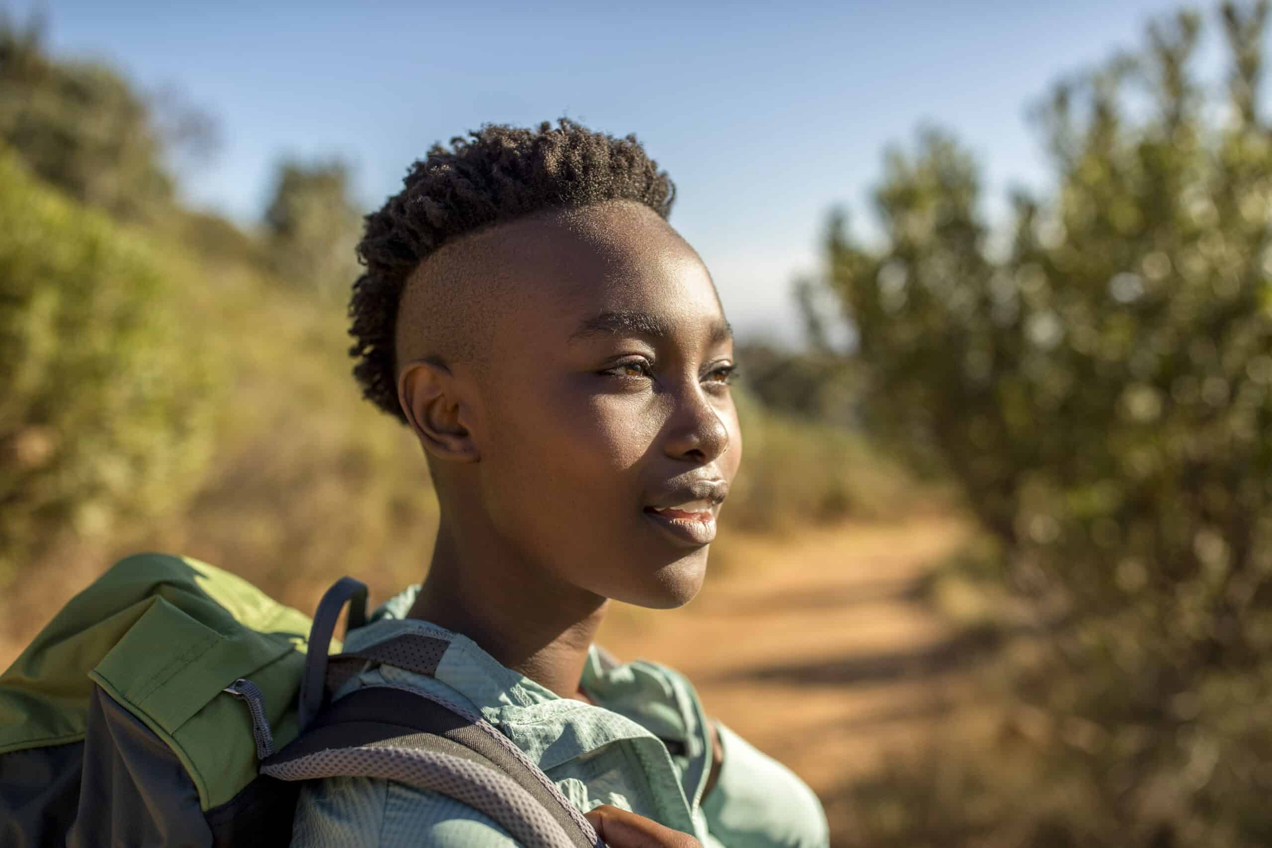 Eine junge schwarze Frau mit Kurzhaarschnitt blickt selbstbewusst nach vorne, während sie einen Rucksack trägt und von sonnenbeschienenem Grün umgeben ist. © Fotografie Tomas Rodriguez