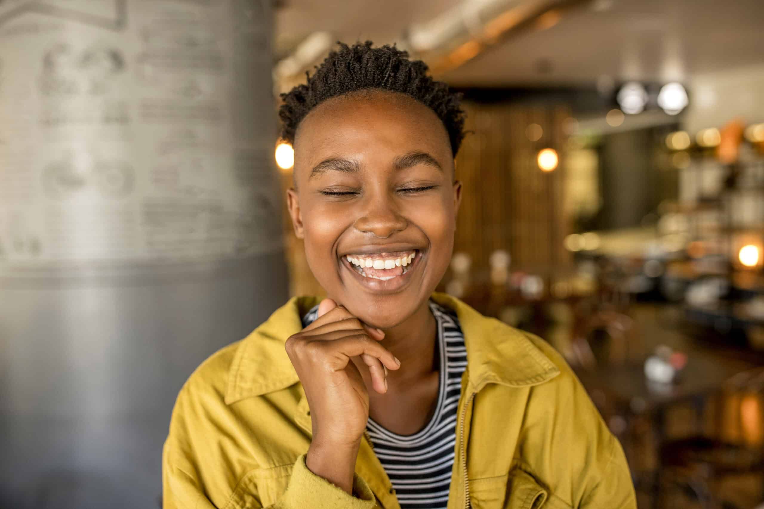 Eine fröhliche junge schwarze Frau mit kurzen Haaren, eine gelbe Jacke und ein gestreiftes Oberteil tragend, lacht mit geschlossenen Augen und der Hand am Kinn in einem warm beleuchteten Café. © Fotografie Tomas Rodriguez