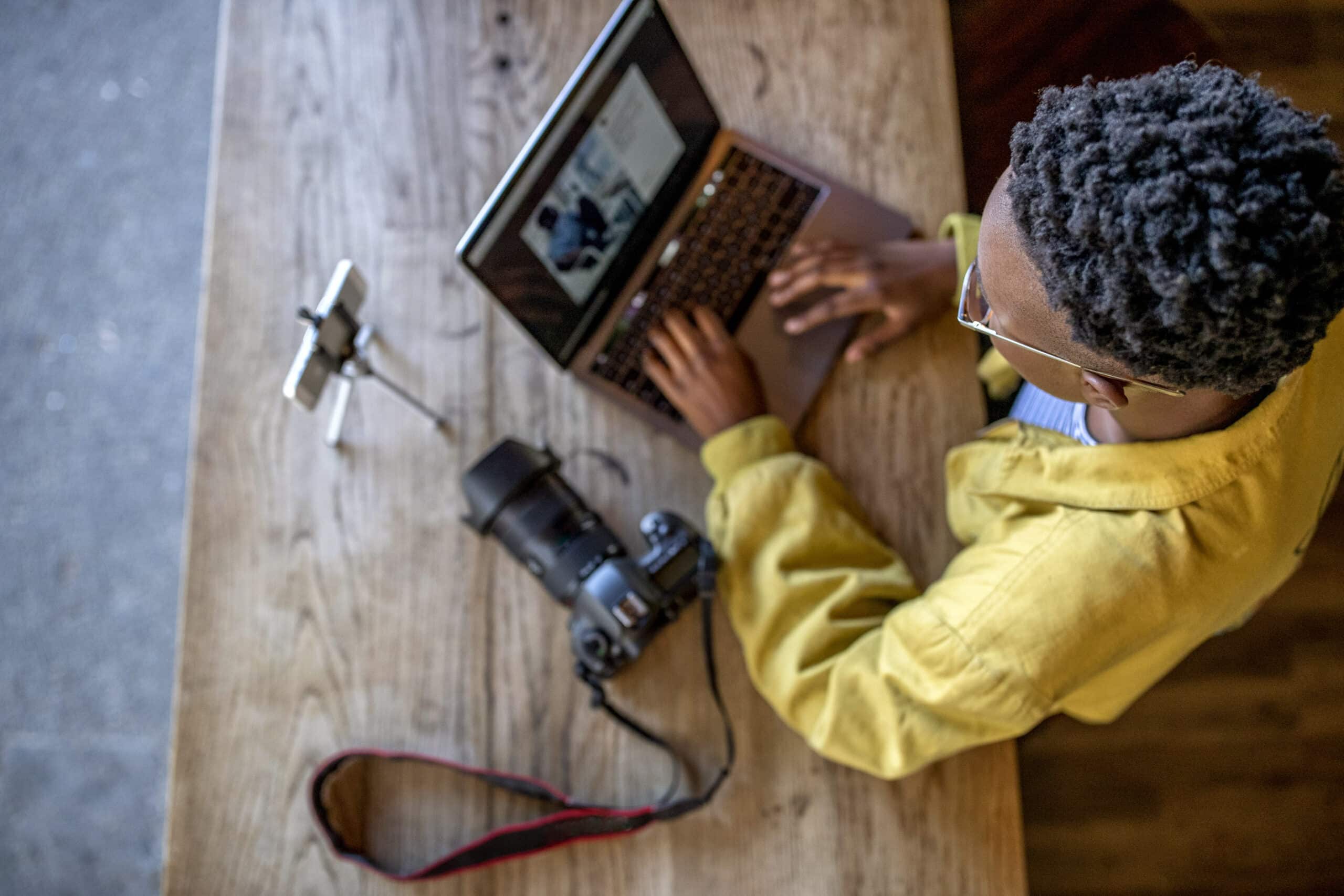 Eine Draufsicht auf eine Person mit kurzen Haaren, Brille und gelbem Hemd, die an einem Holztisch auf einem Laptop tippt, neben ihr steht eine Kamera. © Fotografie Tomas Rodriguez