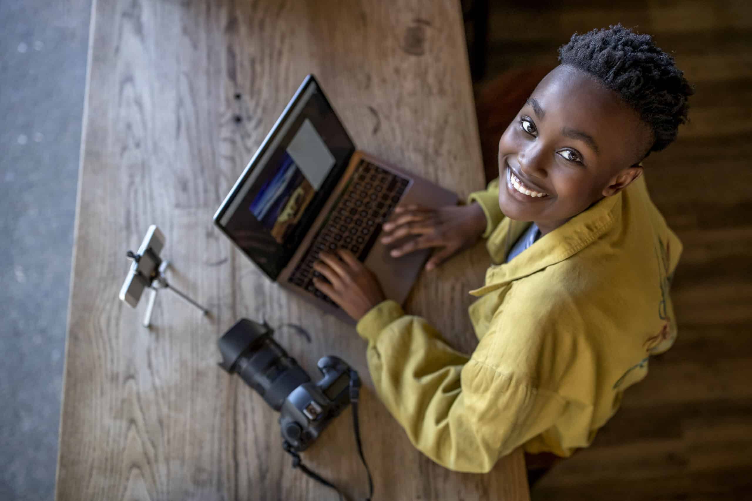 Eine junge Frau mit kurzen Haaren und einem gelben Hemd lächelt, während sie an einem Holztisch mit einer Kamera neben sich einen Laptop benutzt. © Fotografie Tomas Rodriguez