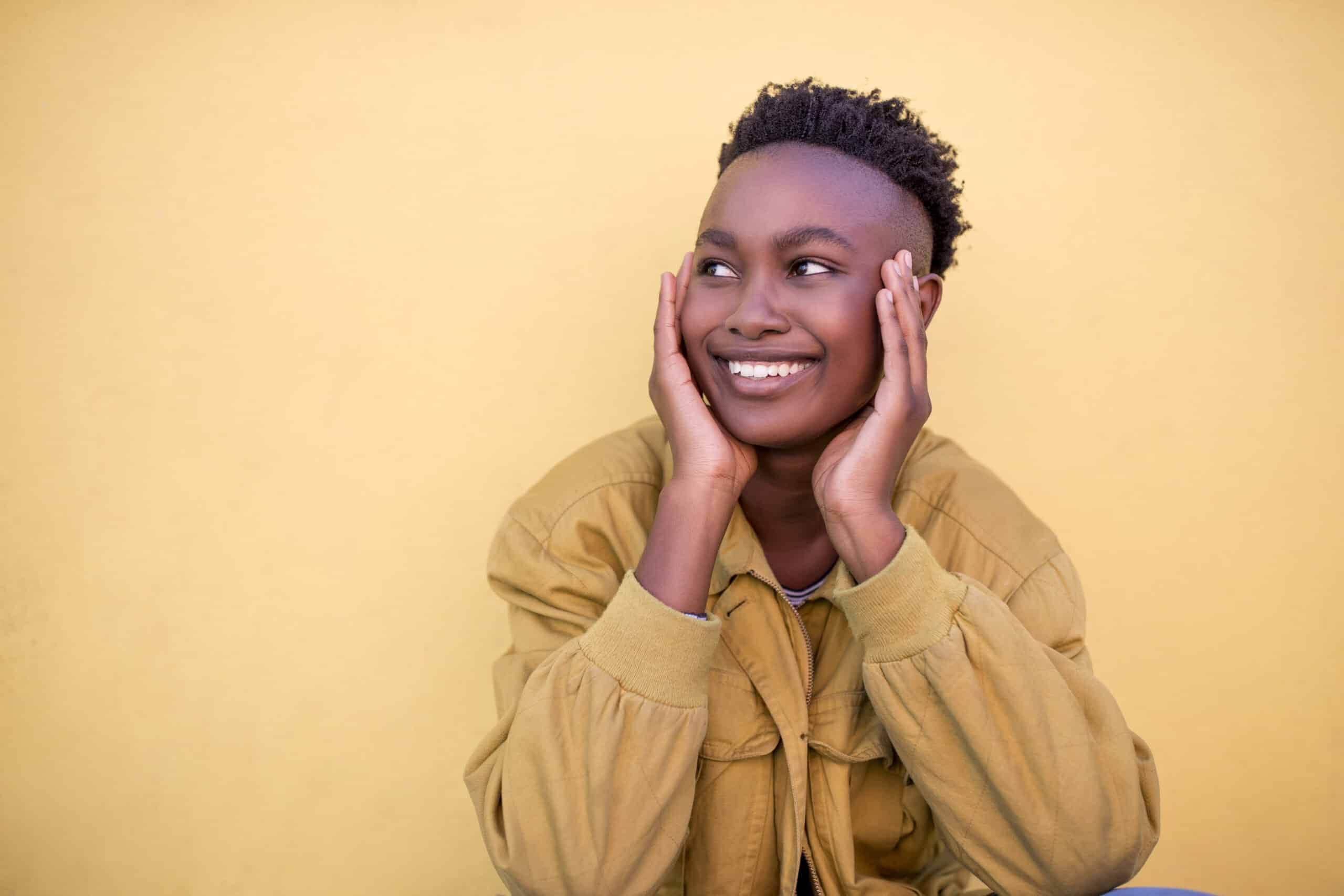 Eine fröhliche junge schwarze Frau mit Kurzhaarfrisur und hellbrauner Jacke, die lächelt, während sie nach oben schaut und ihre Wangen auf ihren Händen abstützt, vor einem sanften gelben Hintergrund. © Fotografie Tomas Rodriguez
