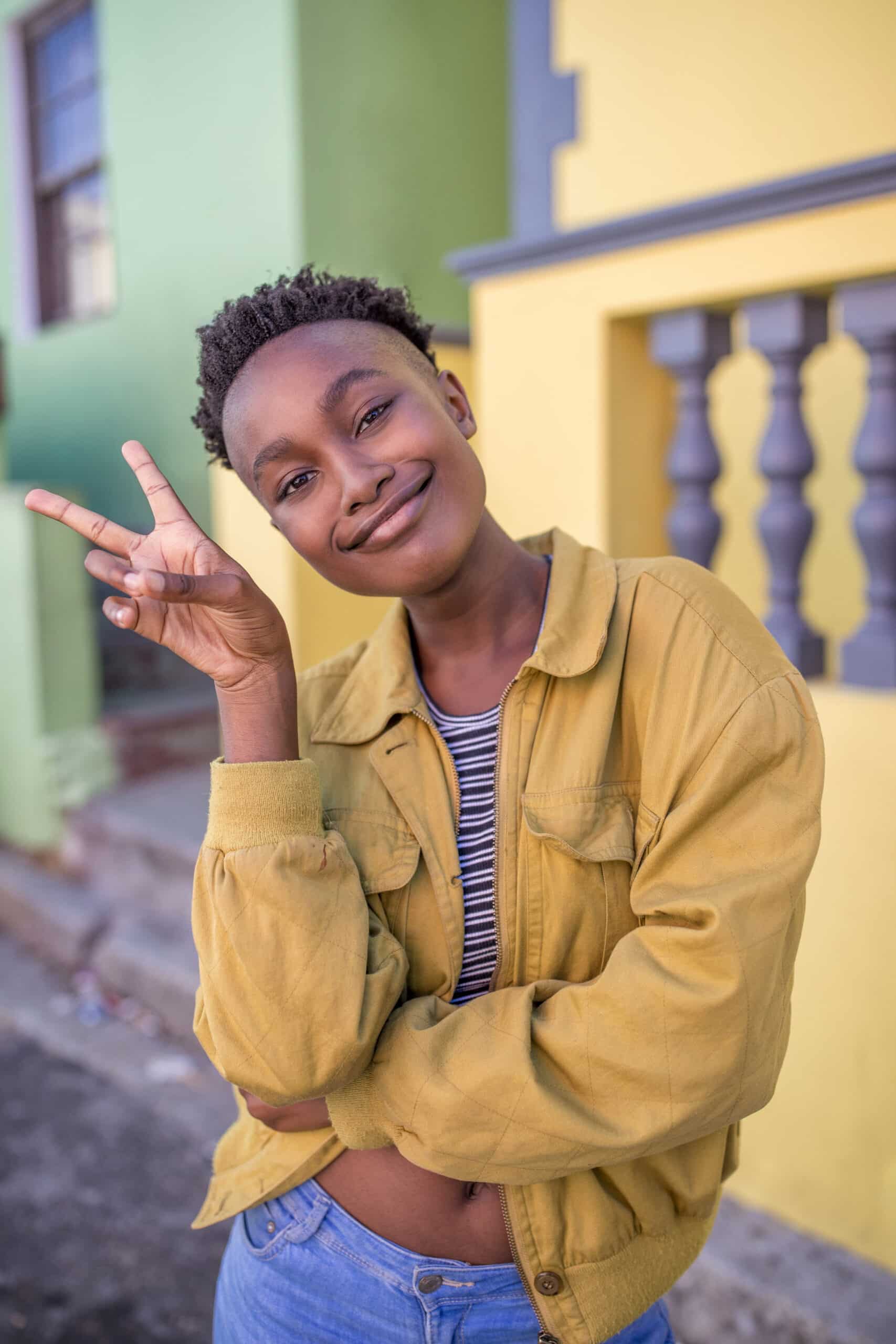 Eine fröhliche junge Frau mit kurzen Haaren lächelt in die Kamera und macht ein Peace-Zeichen, während sie an einer bunten Wand lehnt. Sie trägt eine gelbe Jacke über einem gestreiften Oberteil und blaue Jeans. © Fotografie Tomas Rodriguez