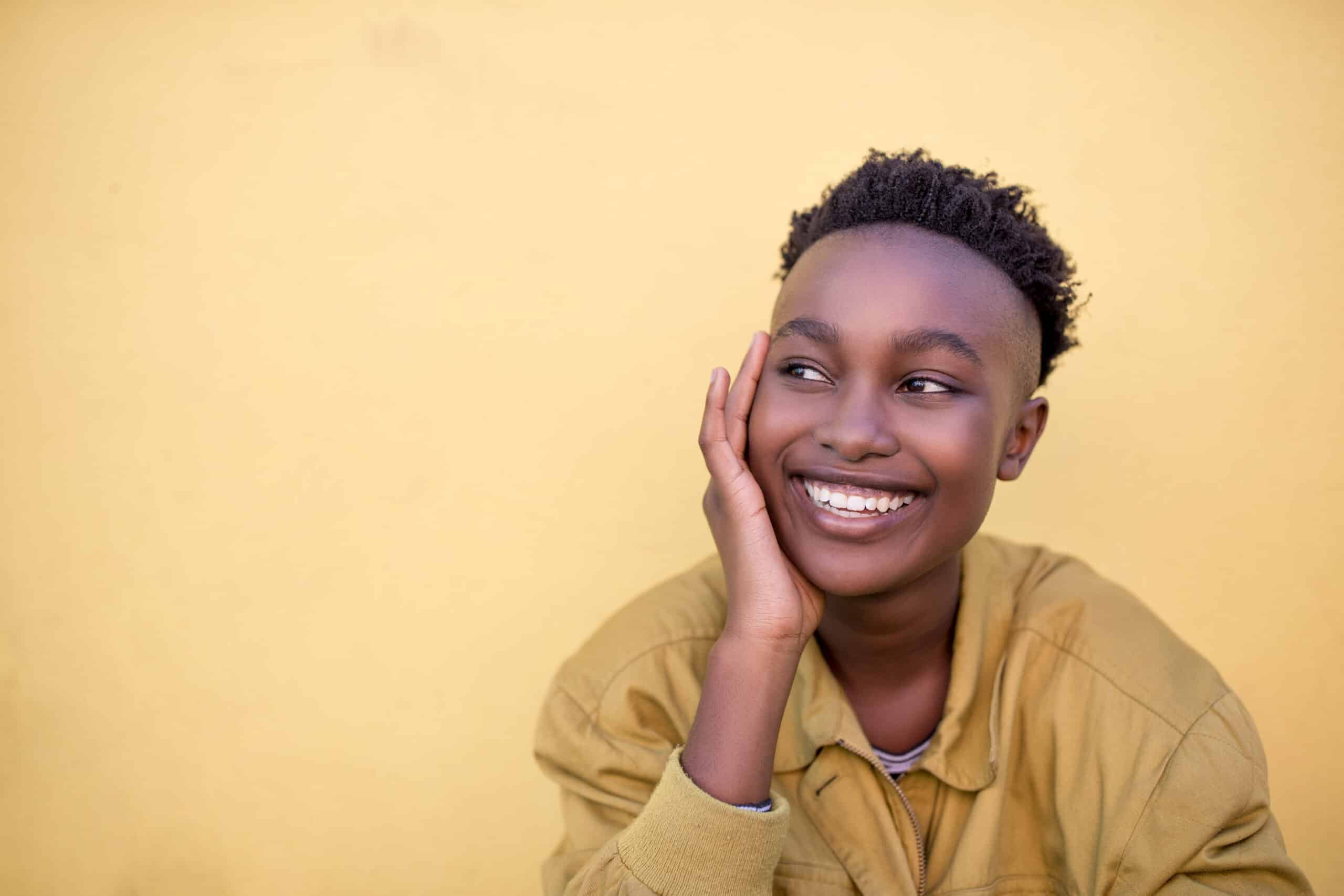 Junge schwarze Frau mit freudigem Gesichtsausdruck, die ihr Kinn auf die Hand stützt und vor einem gelben Hintergrund wegschaut. Sie trägt eine legere Jacke und hat kurzes, lockiges Haar. © Fotografie Tomas Rodriguez