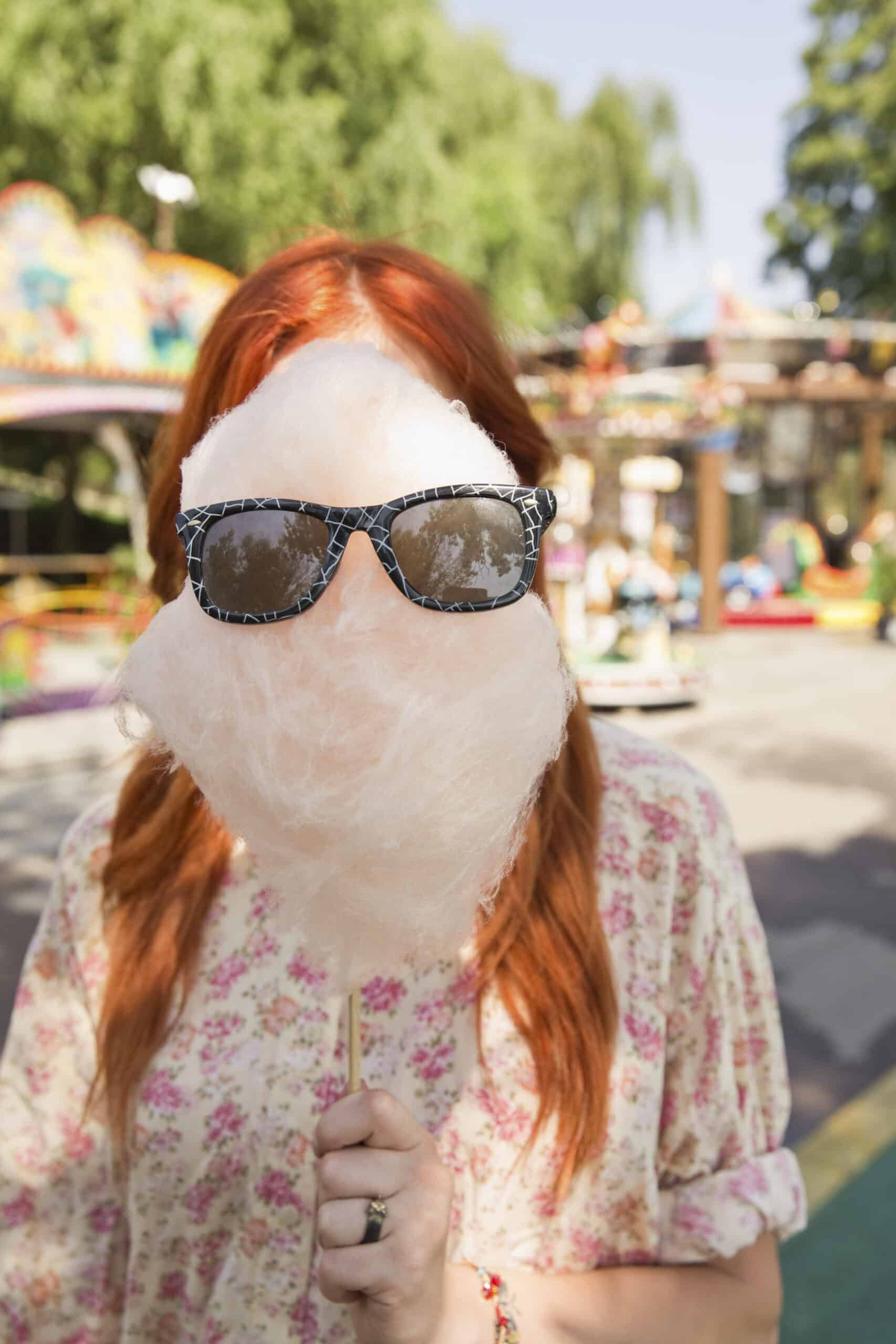 Eine Frau mit roten Haaren hält eine große Zuckerwatte mit einer Sonnenbrille darauf vor ihr Gesicht, auf einem Jahrmarkt mit Fahrgeschäften im Hintergrund. © Fotografie Tomas Rodriguez