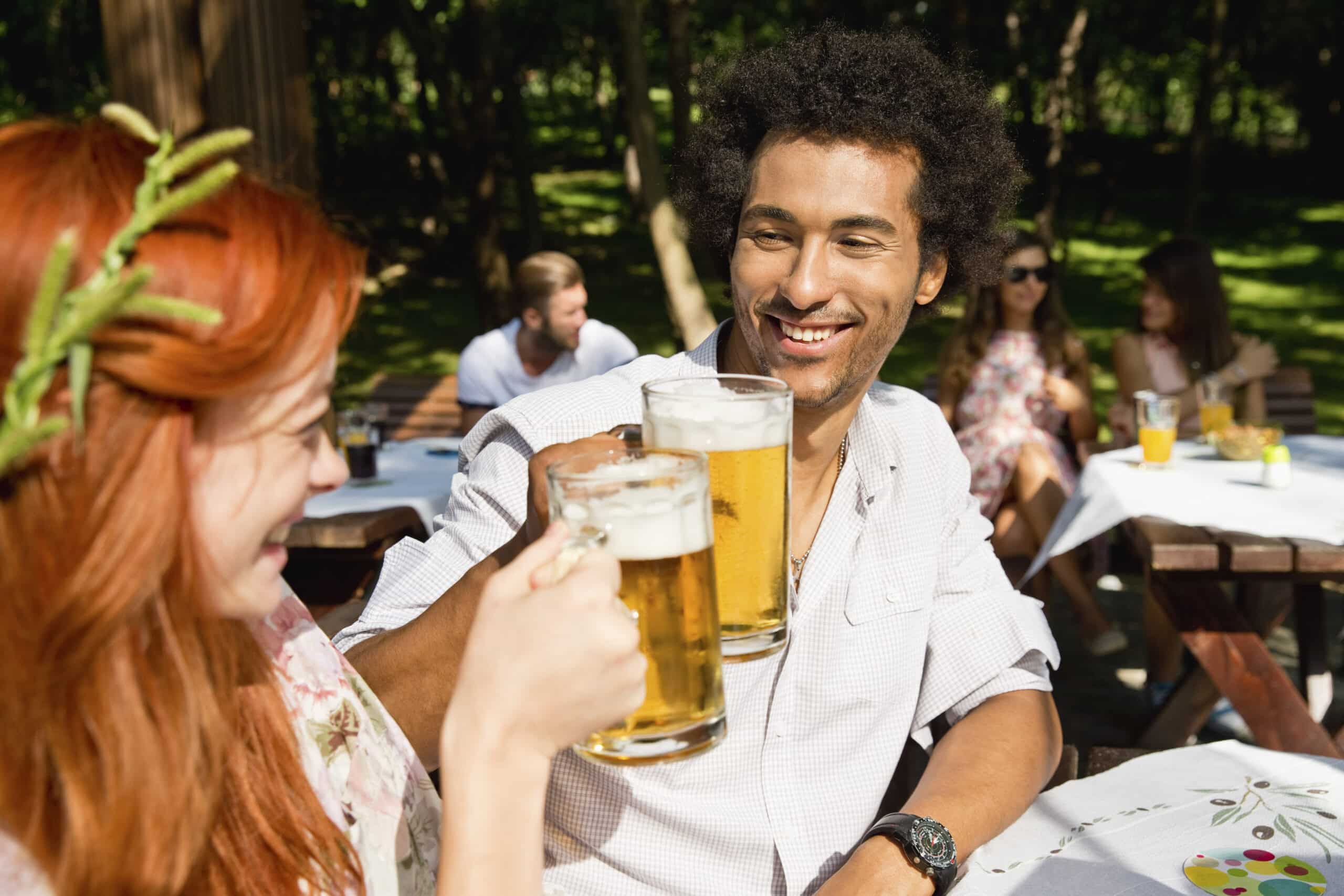 Ein lächelnder junger Mann mit lockigem Haar stößt im Freien mit einer rothaarigen Frau mit Biergläsern an, während sich im sonnenbeschienenen Hintergrund andere unterhalten. © Fotografie Tomas Rodriguez