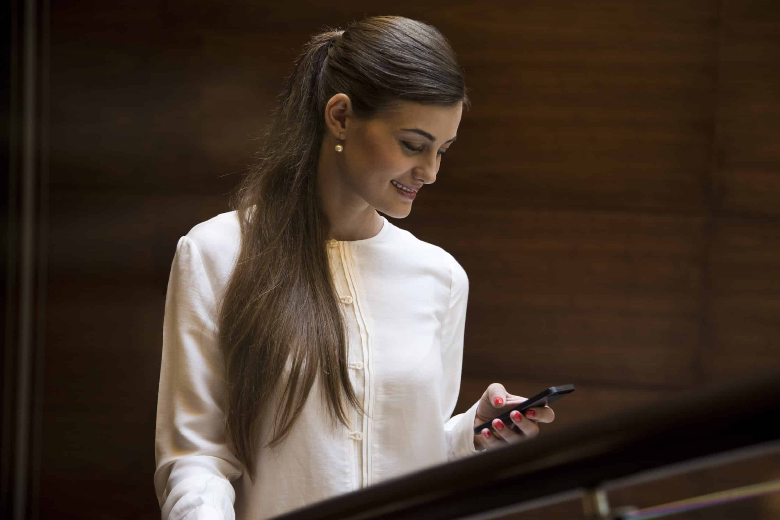 Eine Frau in einer weißen Bluse lächelt, während sie auf ihr Smartphone schaut und in einem Raum mit holzgetäfelten Wänden steht. © Fotografie Tomas Rodriguez