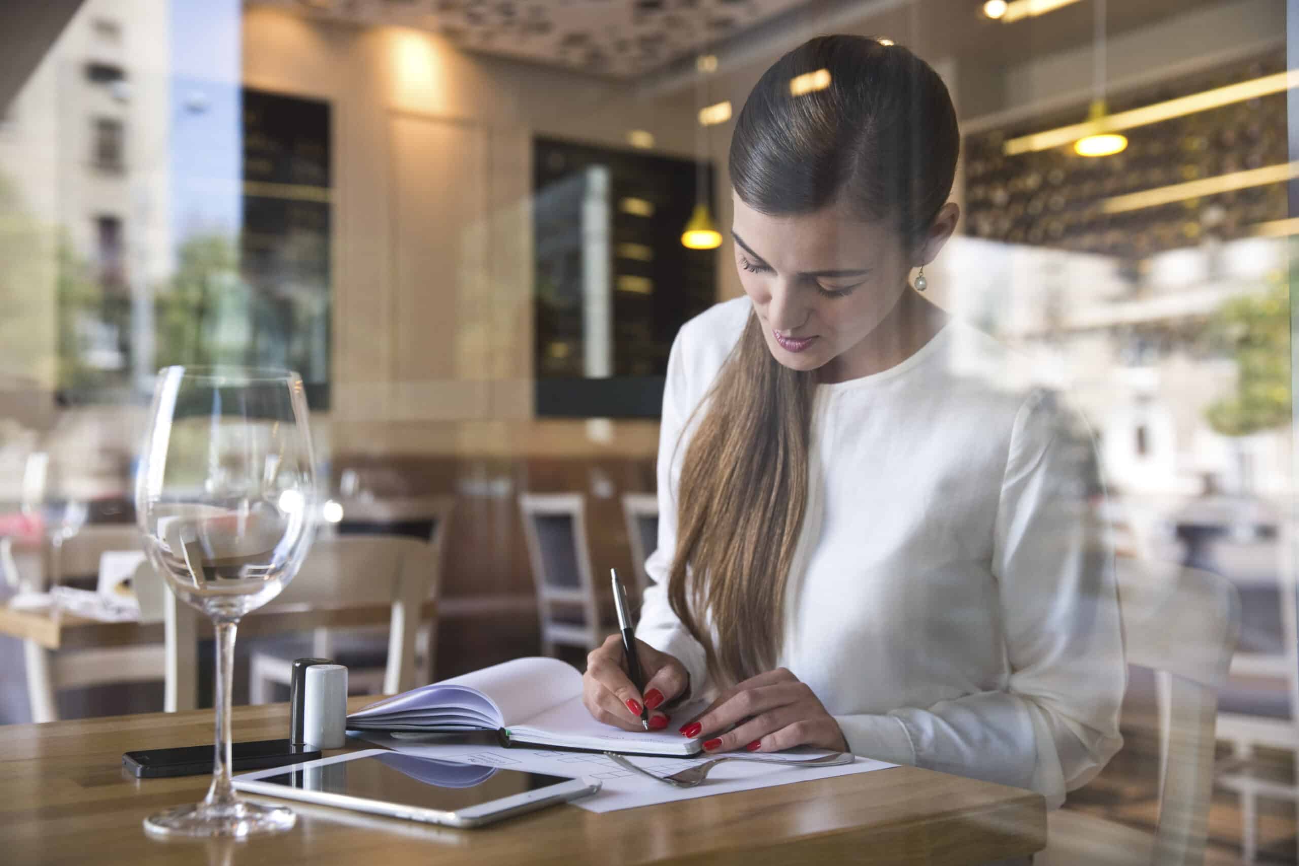Eine Frau in einer weißen Bluse sitzt konzentriert an einem Cafétisch und schreibt in ein Notizbuch. Neben ihr stehen ein Glas Wasser und ein Smartphone. Der Blick ist durch ein Fenster möglich. © Fotografie Tomas Rodriguez