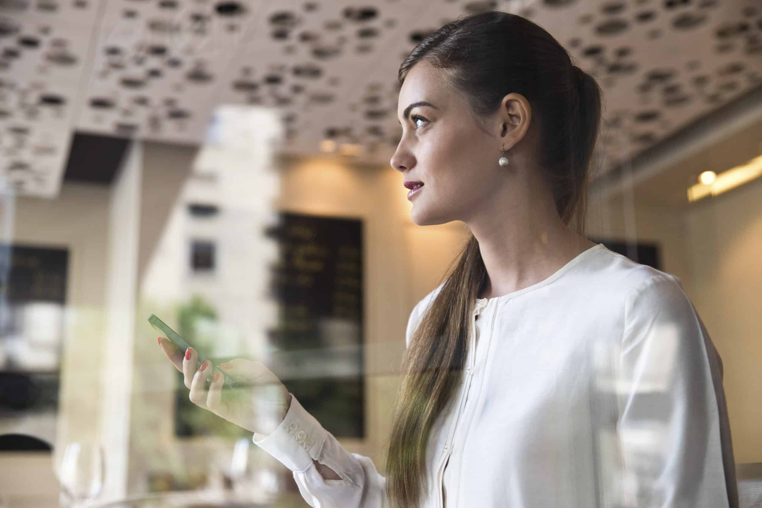 Eine Frau in einer weißen Bluse hält ein Smartphone und blickt durch ein Fenster in einem hell erleuchteten modernen Café mit einzigartigem Deckendekor. © Fotografie Tomas Rodriguez