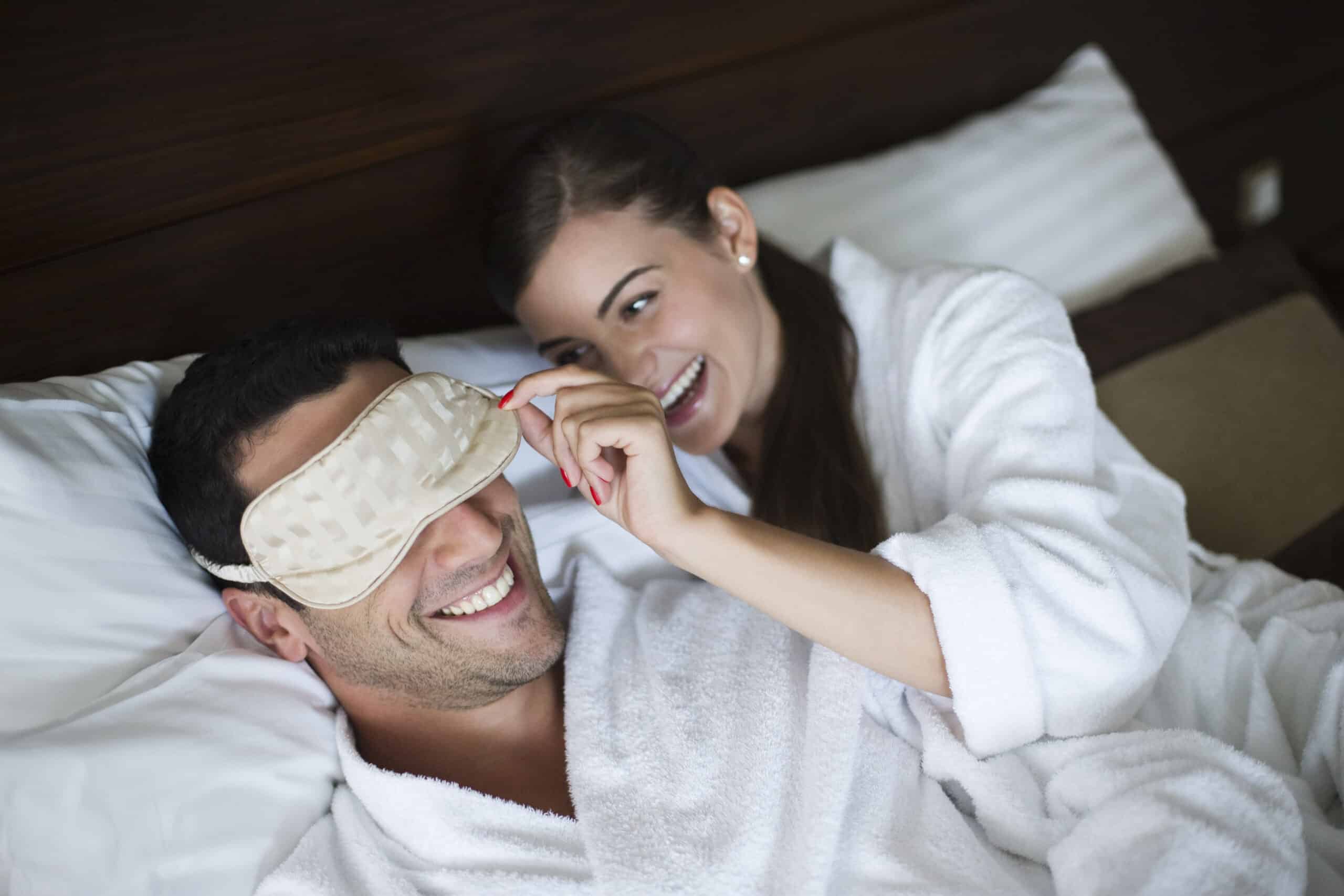 Ein Mann trägt eine Schlafmaske und eine Frau im weißen Bademantel hebt die Maske spielerisch an. Beide lächeln und liegen auf einem Bett. © Fotografie Tomas Rodriguez