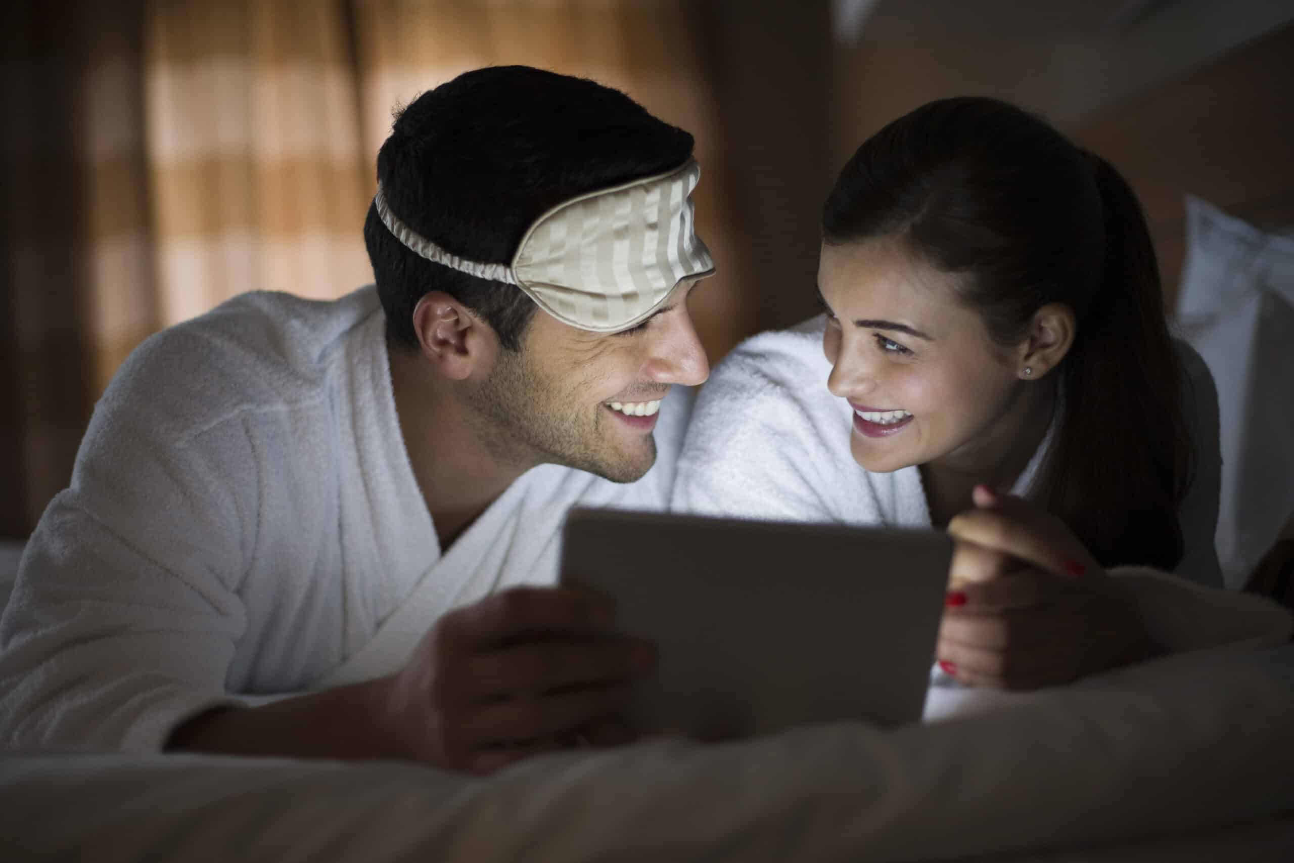 Ein lächelnder Mann und eine lächelnde Frau in weißen Bademänteln liegen im Bett und schauen auf ein Tablet. Der Mann trägt eine Augenmaske, die er auf die Stirn geschoben hat. Das Zimmer ist schwach beleuchtet, was für eine gemütliche Atmosphäre sorgt. © Fotografie Tomas Rodriguez