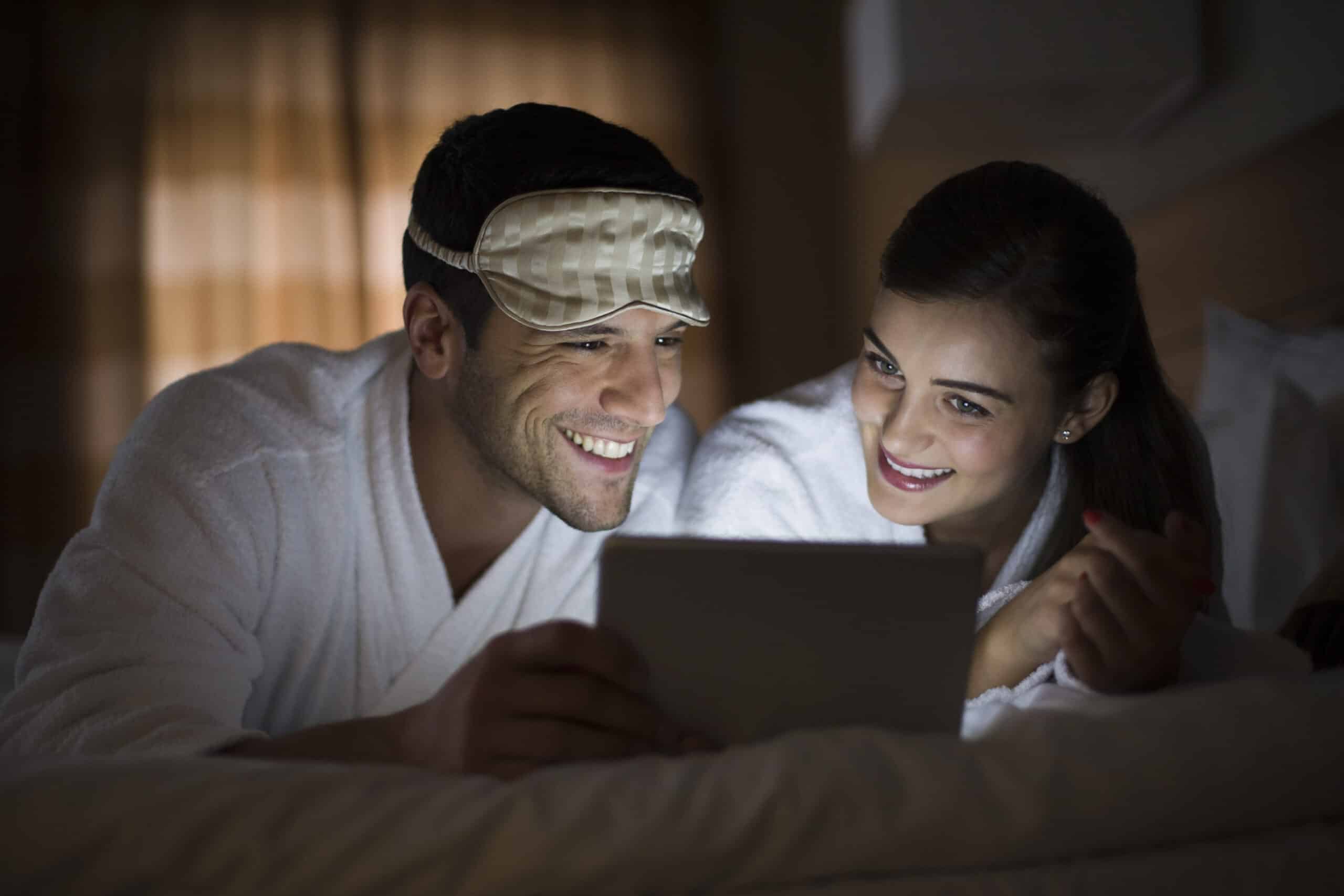 Ein lächelndes Paar in weißen Gewändern schaut in einem schwach beleuchteten Raum auf ein Tablet, wobei der Mann eine auf die Stirn geschobene Augenmaske trägt. © Fotografie Tomas Rodriguez